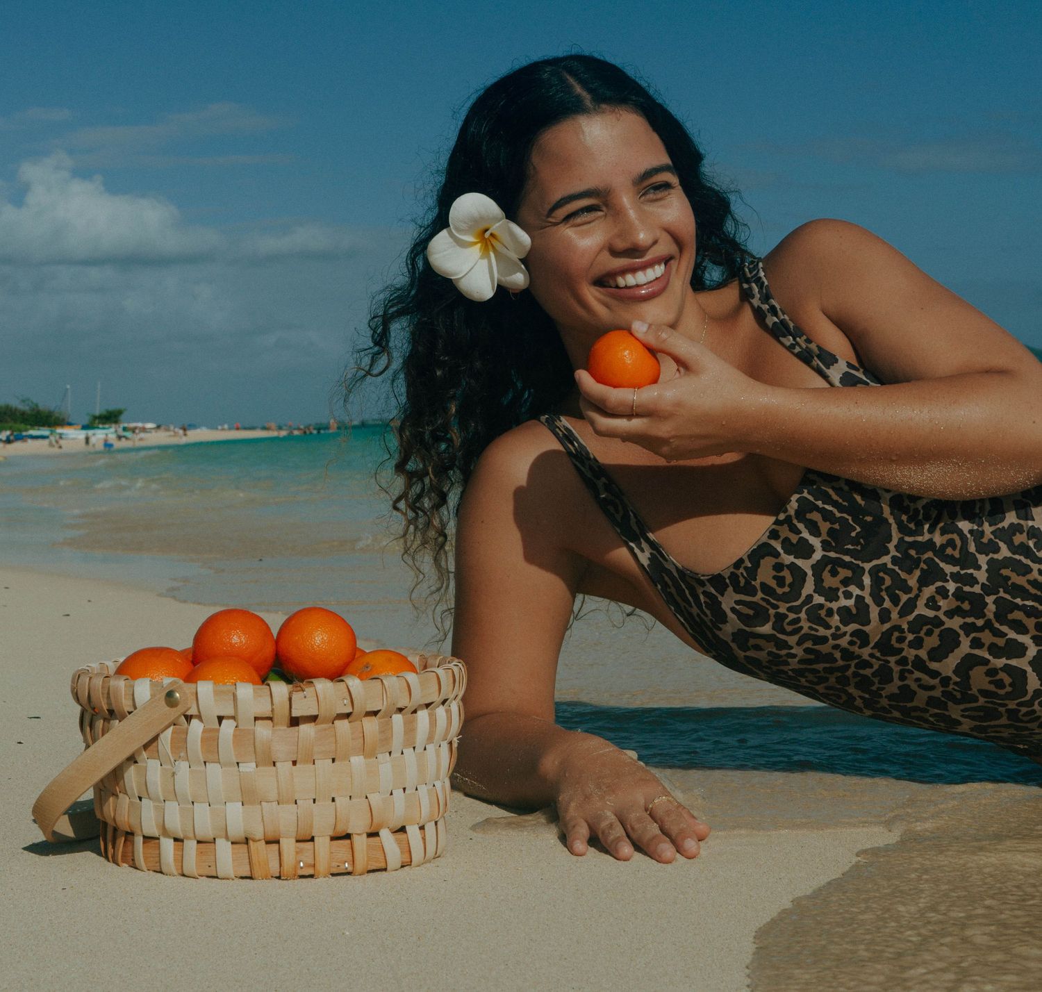 Smiling woman in leopard swimsuit lying on beach holding orange beside wicker basket of oranges.