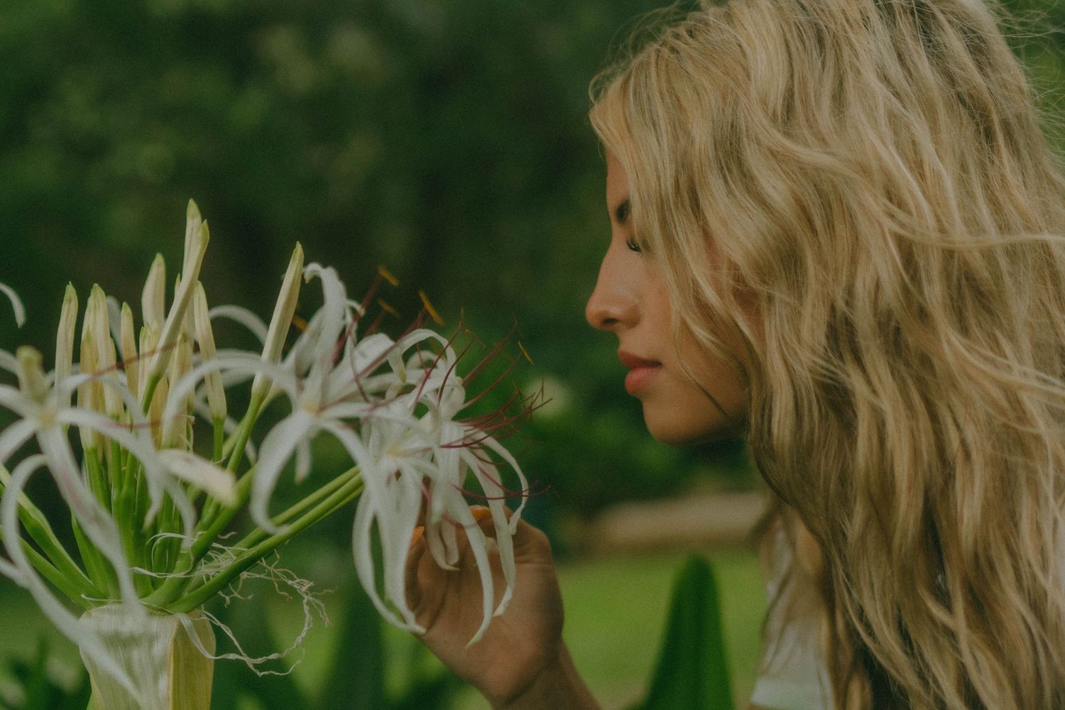 Blonde woman in profile smelling delicate white spider lily flowers in lush garden.