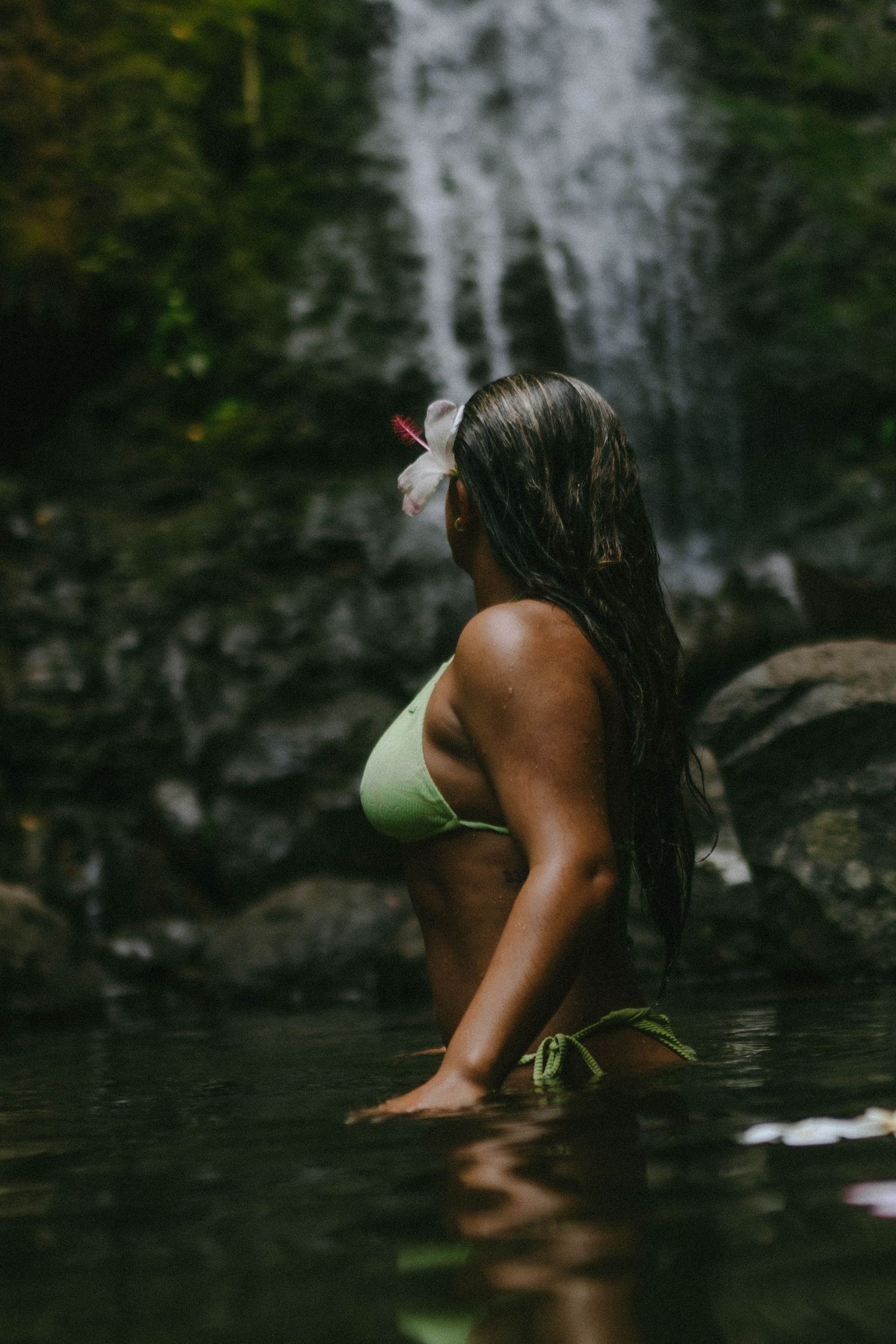 Woman in green bikini sitting in water before a tropical waterfall with hibiscus in hair.