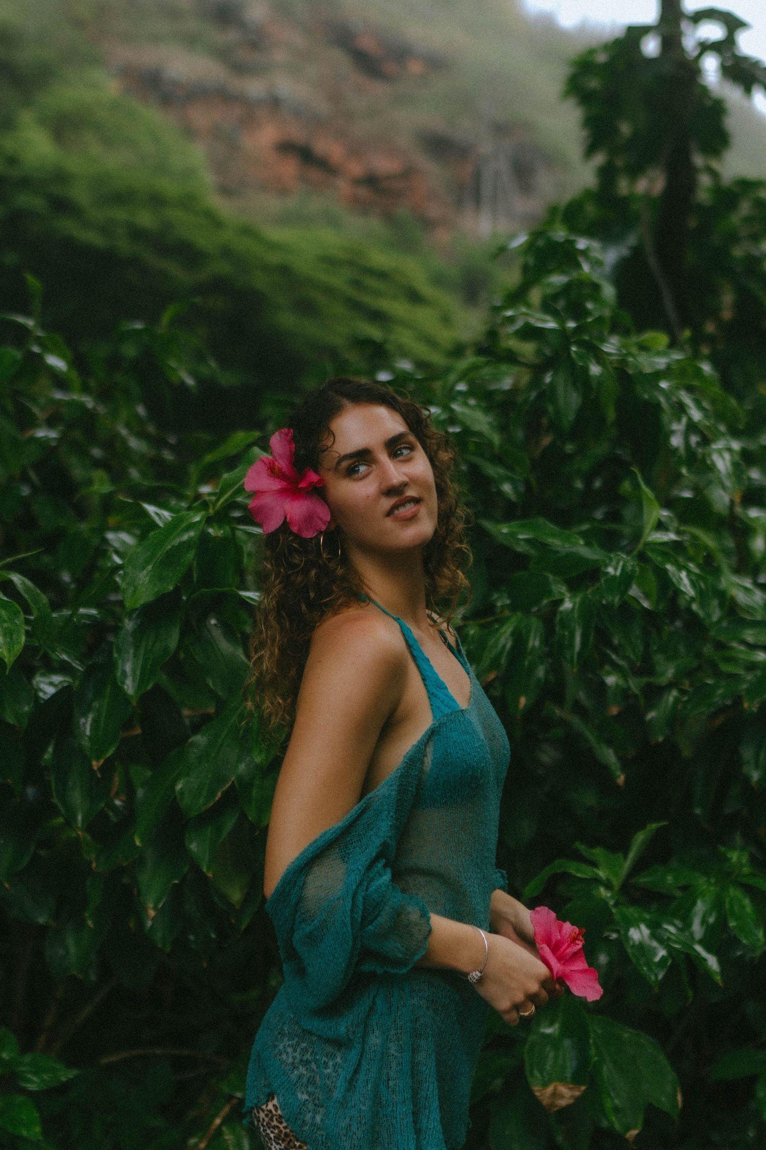 Woman with pink hibiscus flower in curly hair wearing teal dress among tropical foliage.