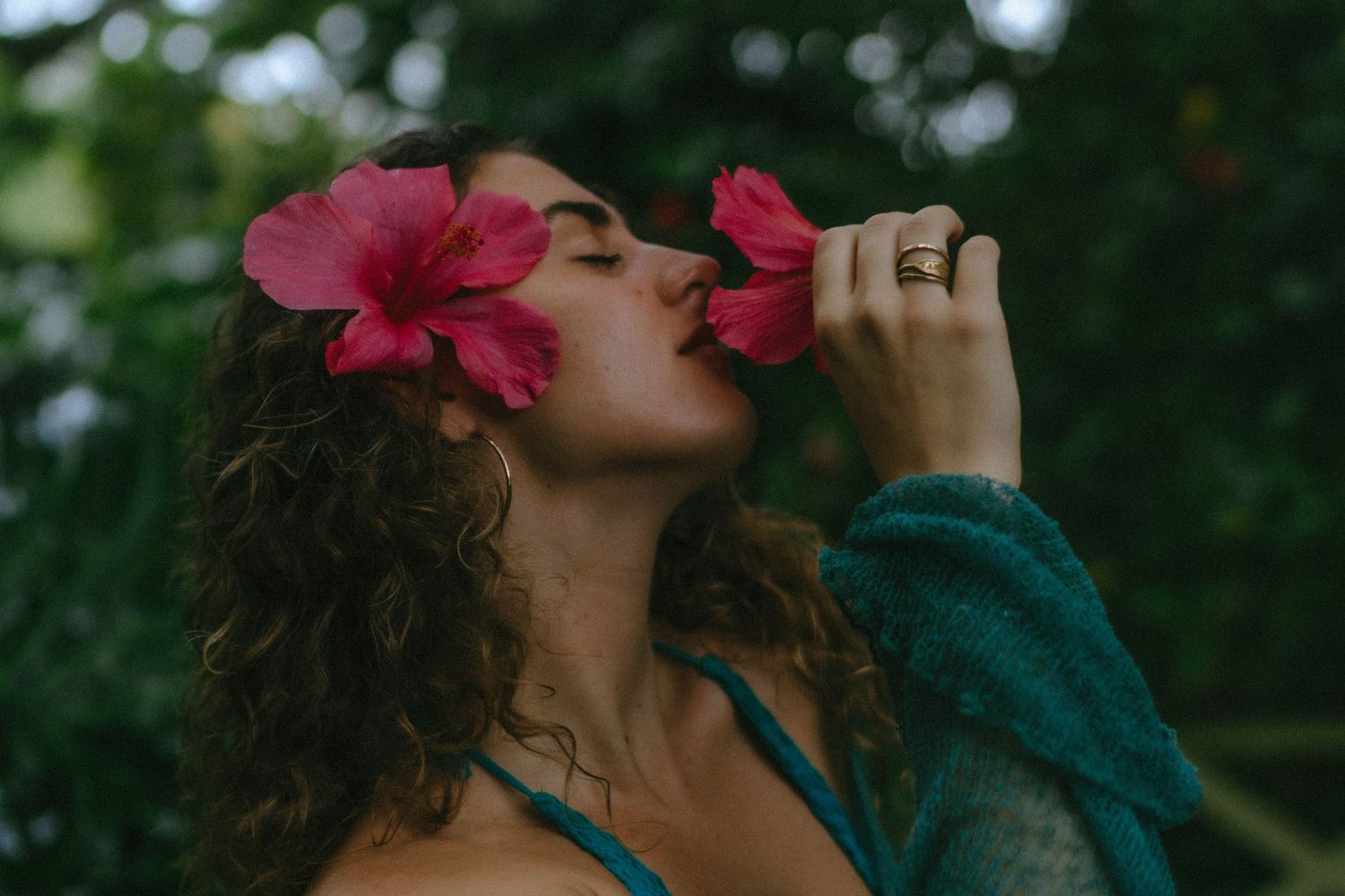 Woman with curly hair smelling pink hibiscus flowers in lush green garden.