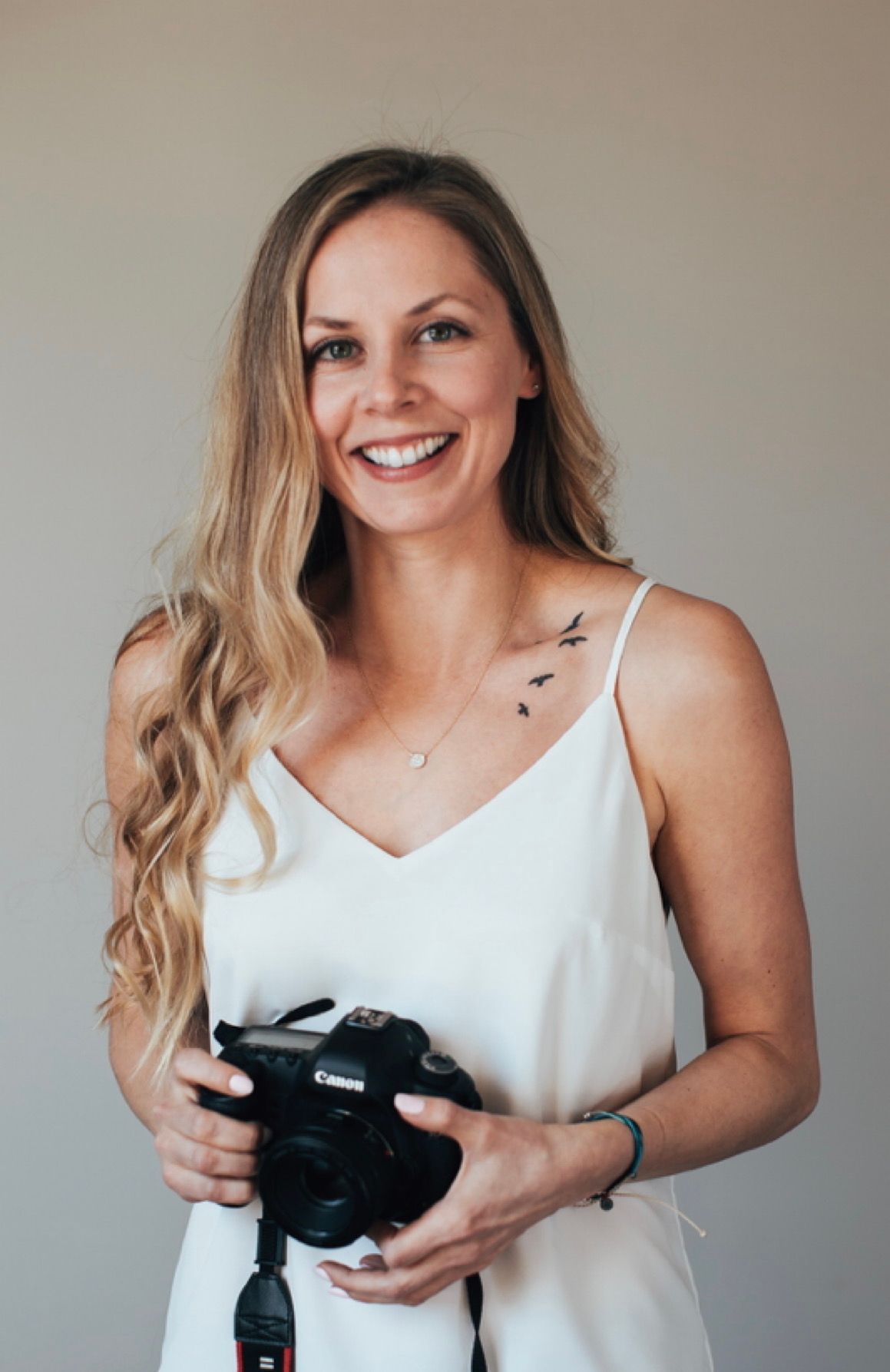 Smiling blonde woman holding a Canon camera, wearing a white dress with bird tattoo on chest.