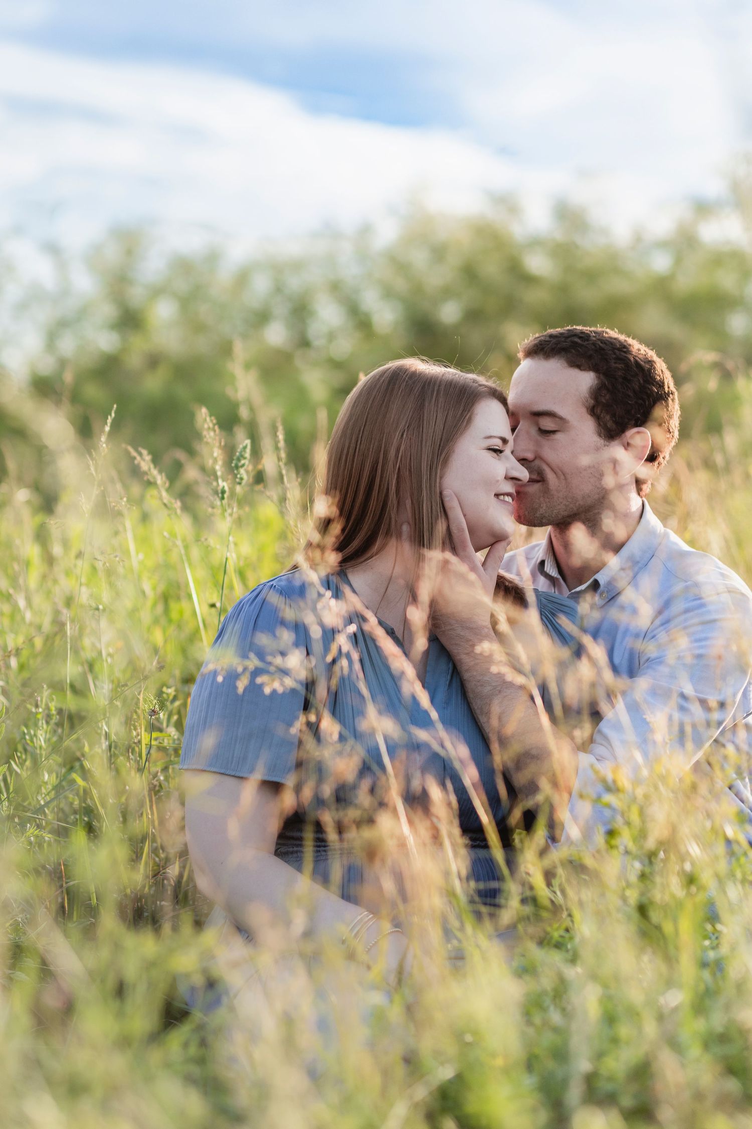 Couple sitting in tall green grass field, sharing a romantic moment on a sunny day with blue sky background.