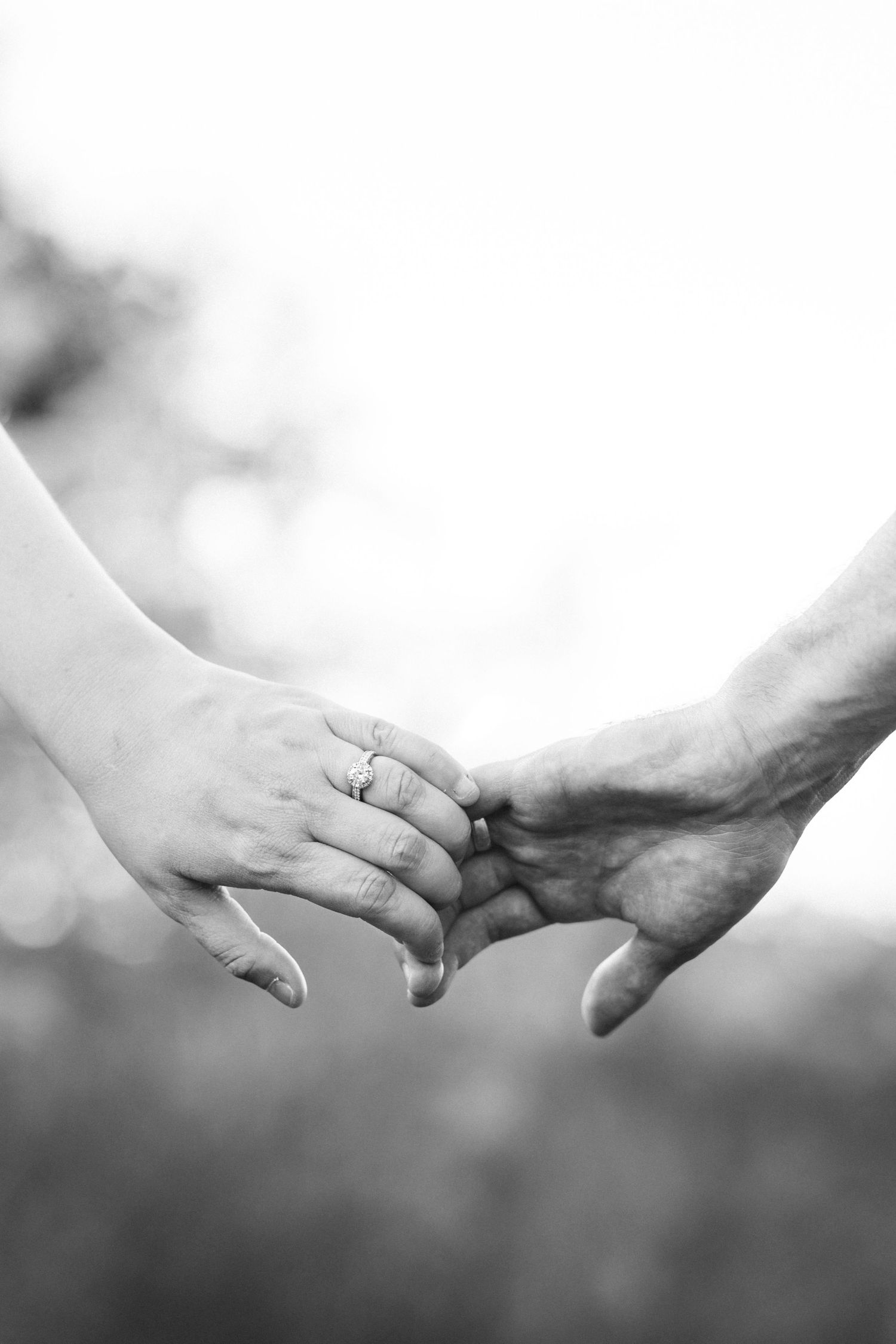 Black and white photo of two hands gently touching, one wearing an engagement ring, symbolizing love and commitment.