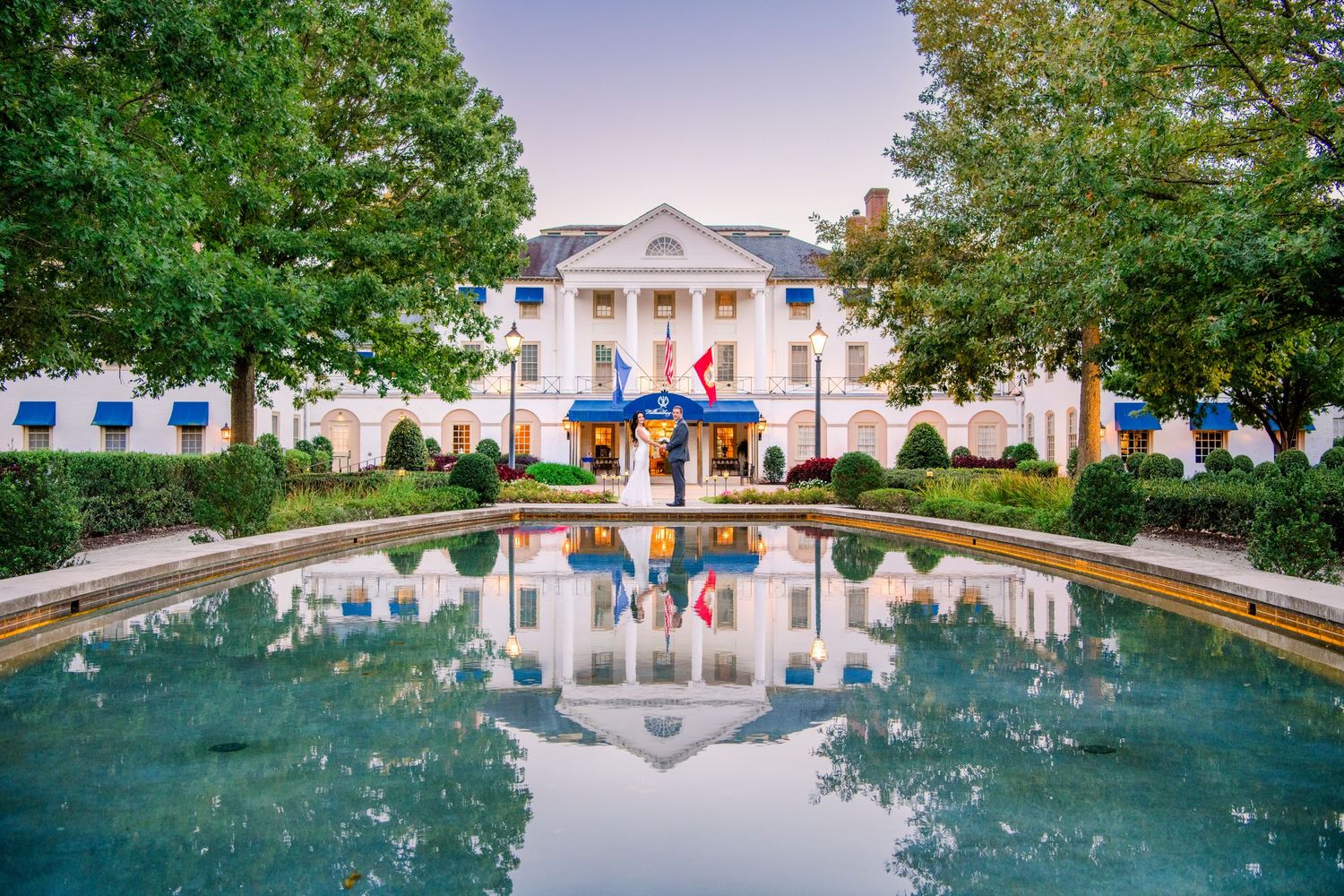 Grand white colonial hotel with blue awnings and flags reflected in a calm pool at dusk, framed by lush green trees.