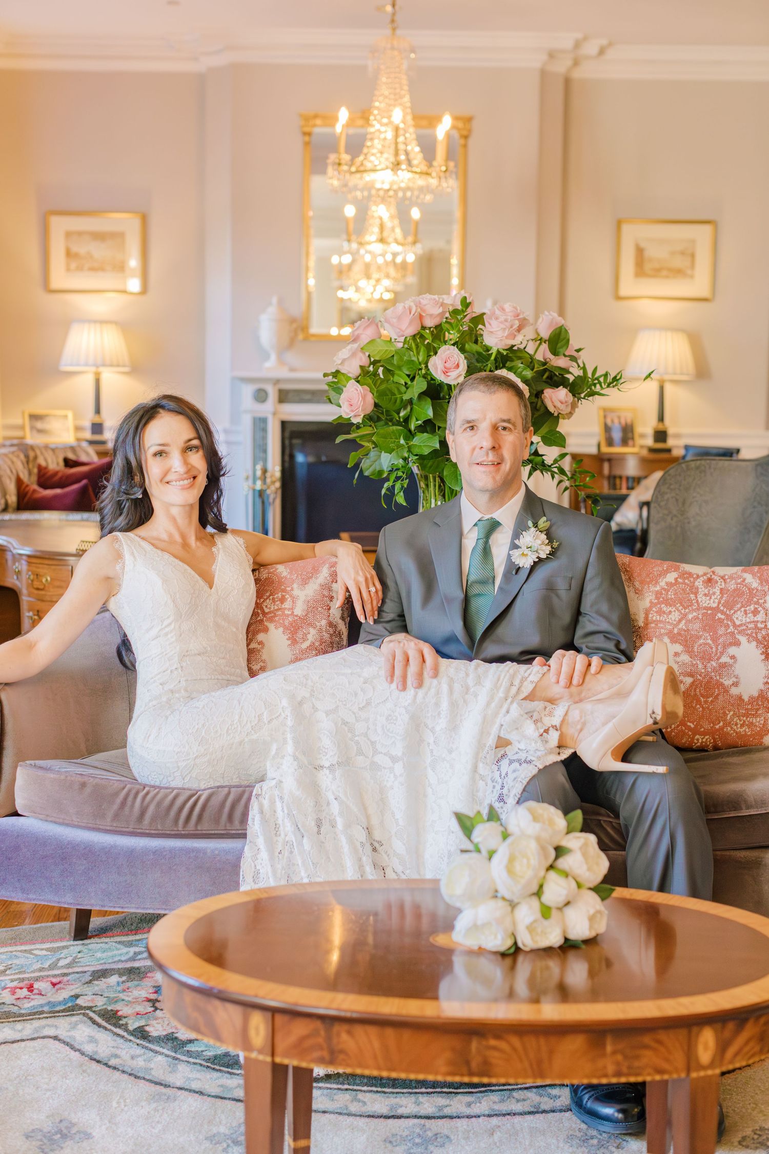 Bride in a white lace dress and groom in a gray suit sit on a sofa in an elegant living room with a white bouquet on the table.
