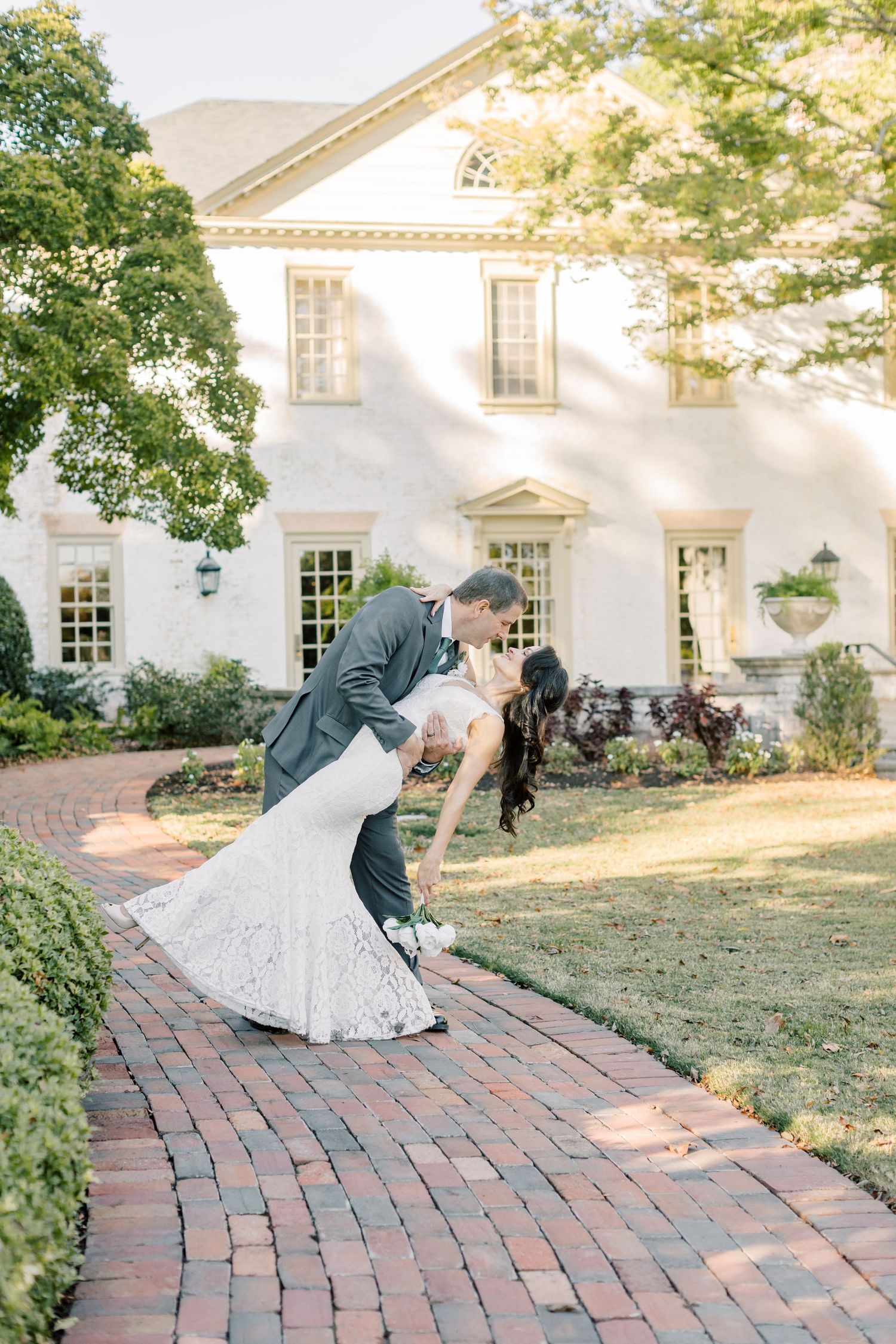 Groom dips bride in a white lace gown on a brick garden path in front of an elegant white historic mansion.