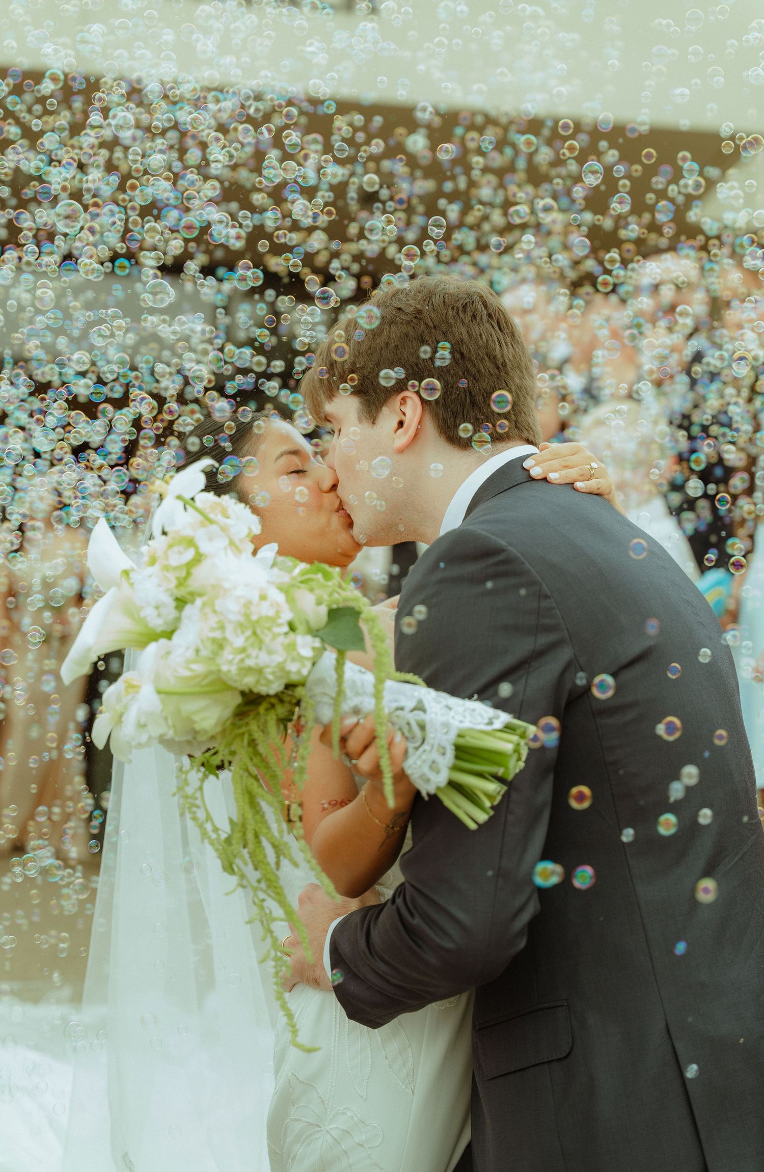 Newlywed couple sharing a romantic kiss surrounded by bubbles, bride holding white floral bouquet in wedding attire.