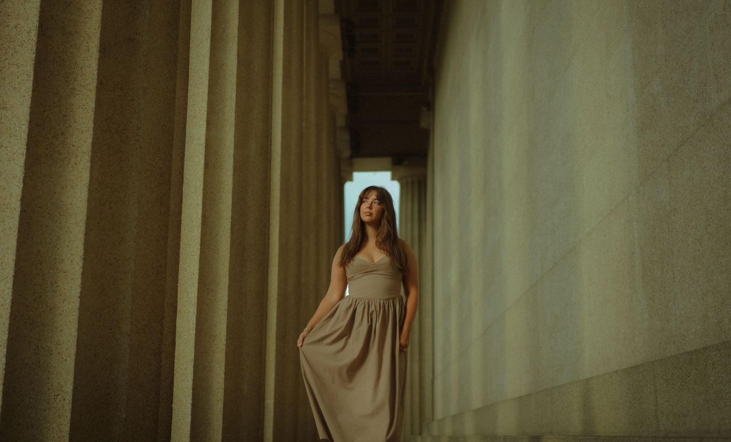 Woman in elegant beige gown standing between tall stone columns in a classical architectural setting.