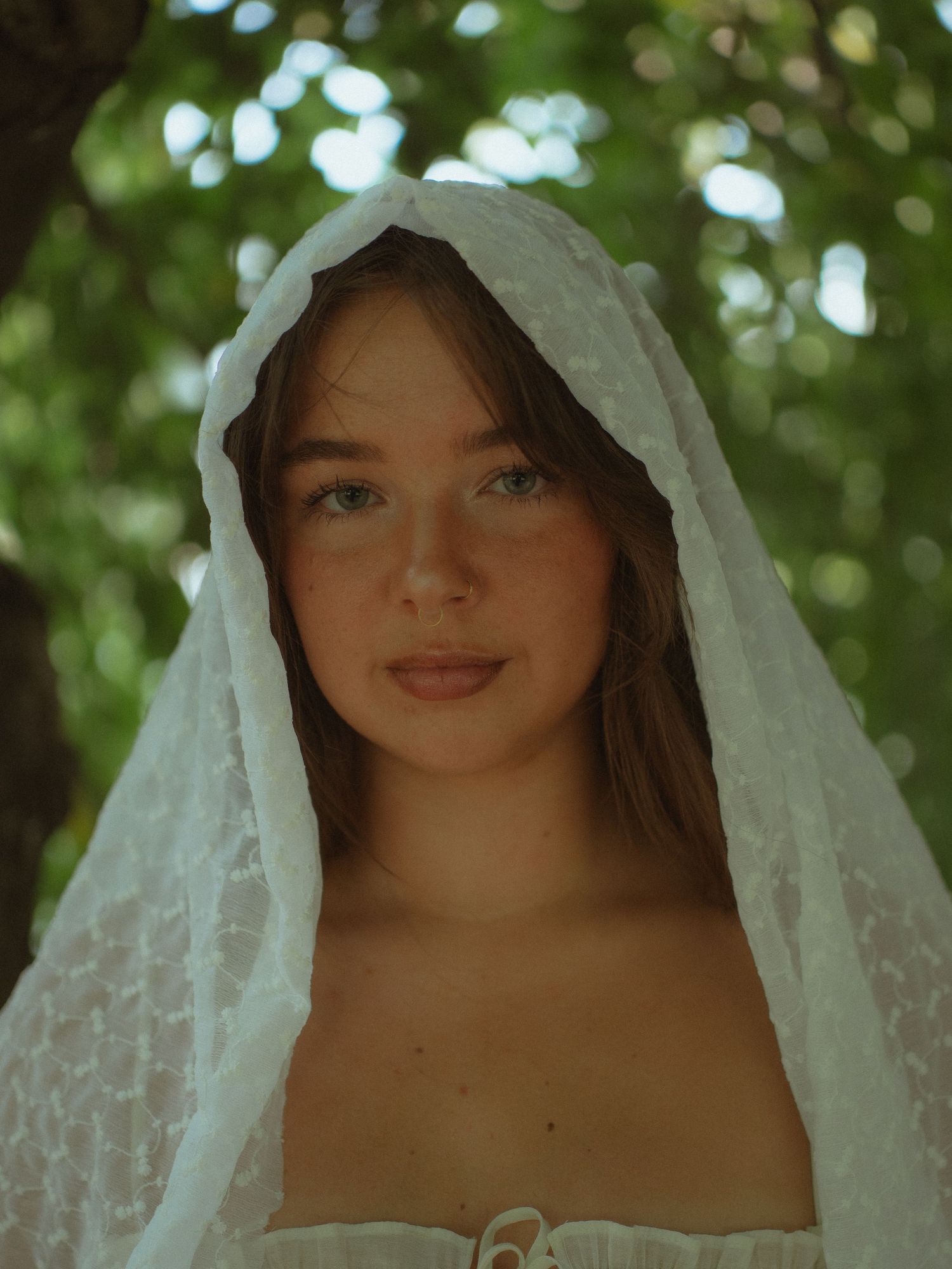 Young woman wearing a white lace veil outdoors with green trees in the background.