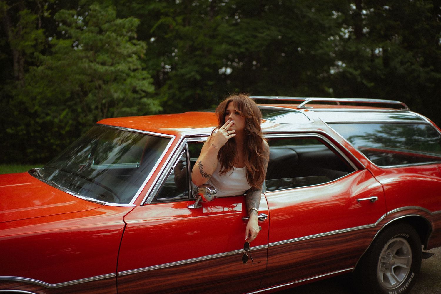 Woman with long hair leaning out of a classic red station wagon window, surrounded by lush green trees.