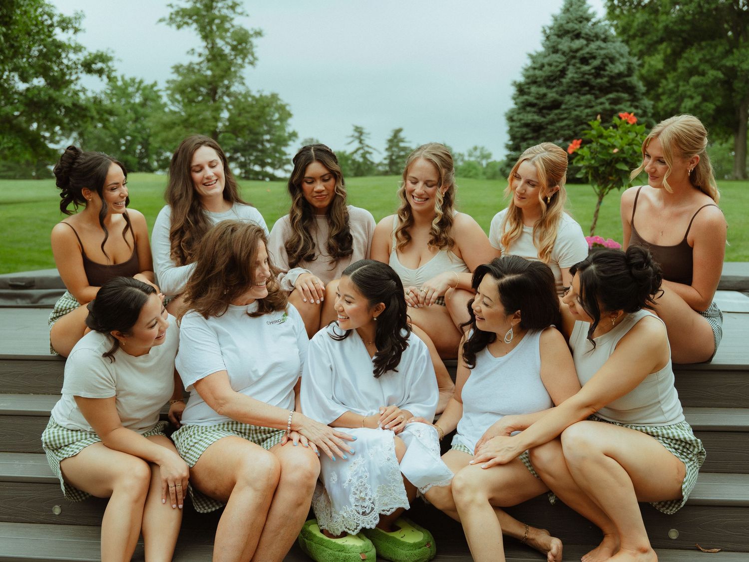 Group of bridesmaids in white and neutral outfits sitting on outdoor steps, laughing and celebrating together.