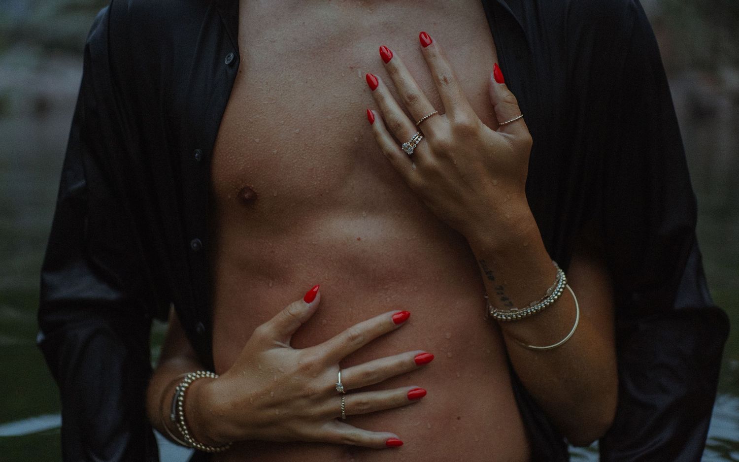 Woman with red nails and gold bracelets placing hands on a man's bare torso, wearing an open black shirt.