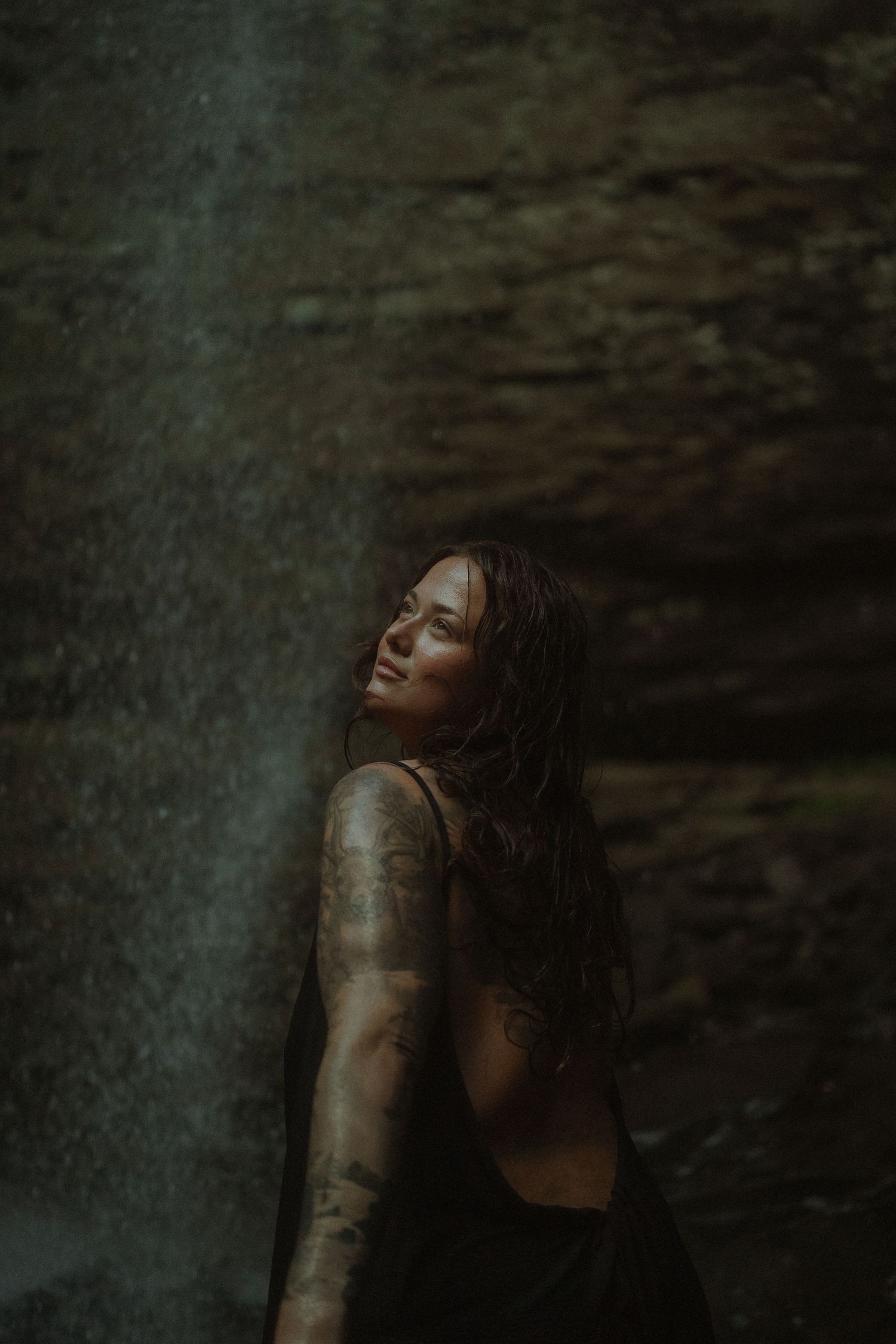 Woman with tattoos standing near a waterfall, head tilted back, eyes closed, wearing a black dress in moody lighting.