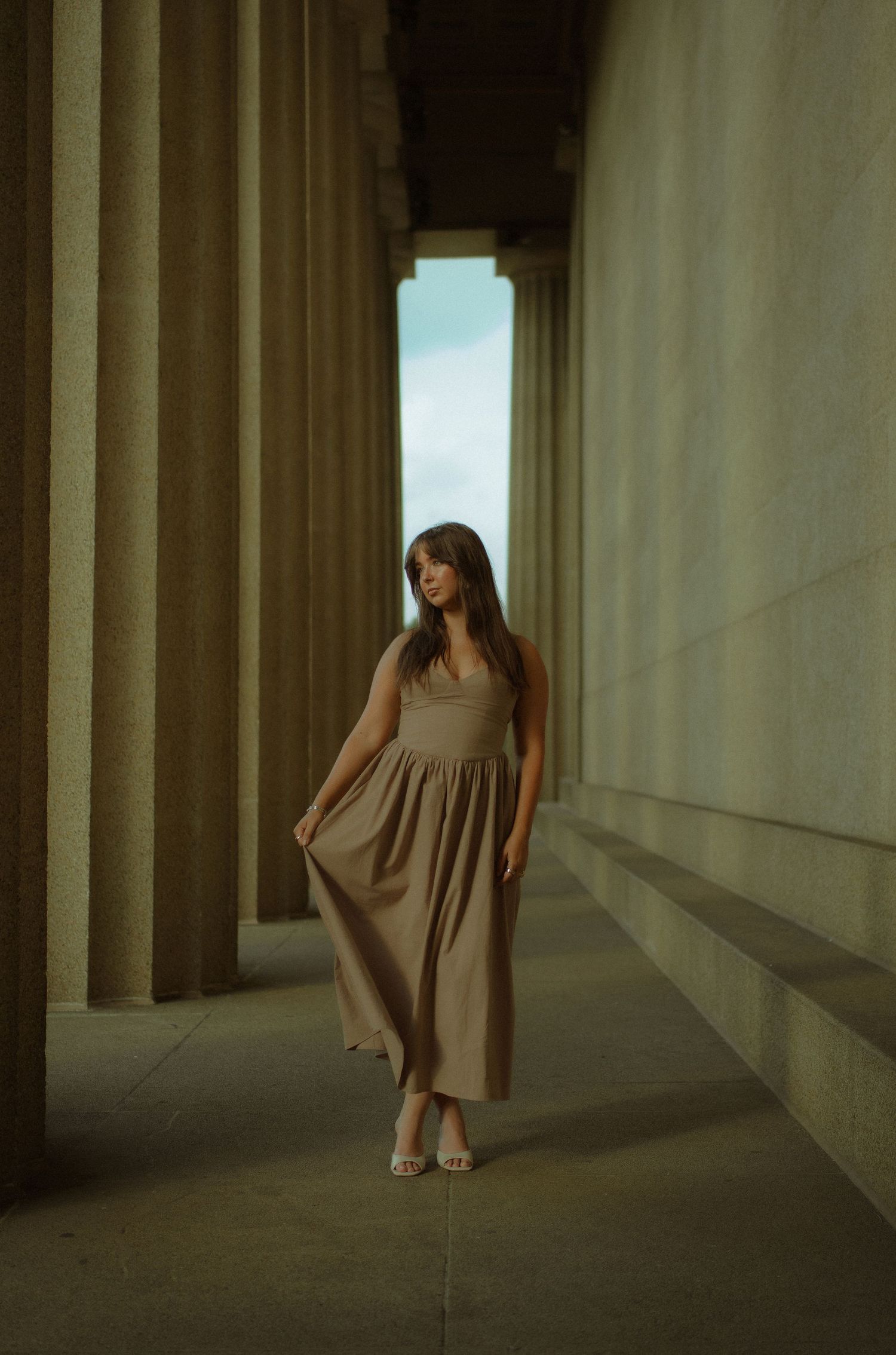 Woman in beige maxi dress posing between large stone columns at a classical architectural monument.