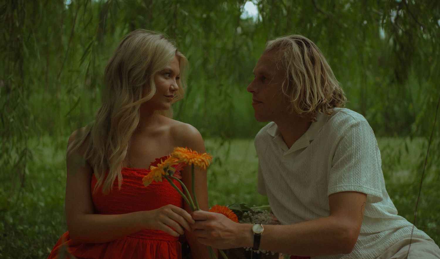 A blonde woman in a red dress receives orange flowers from a man with dreadlocks outdoors.