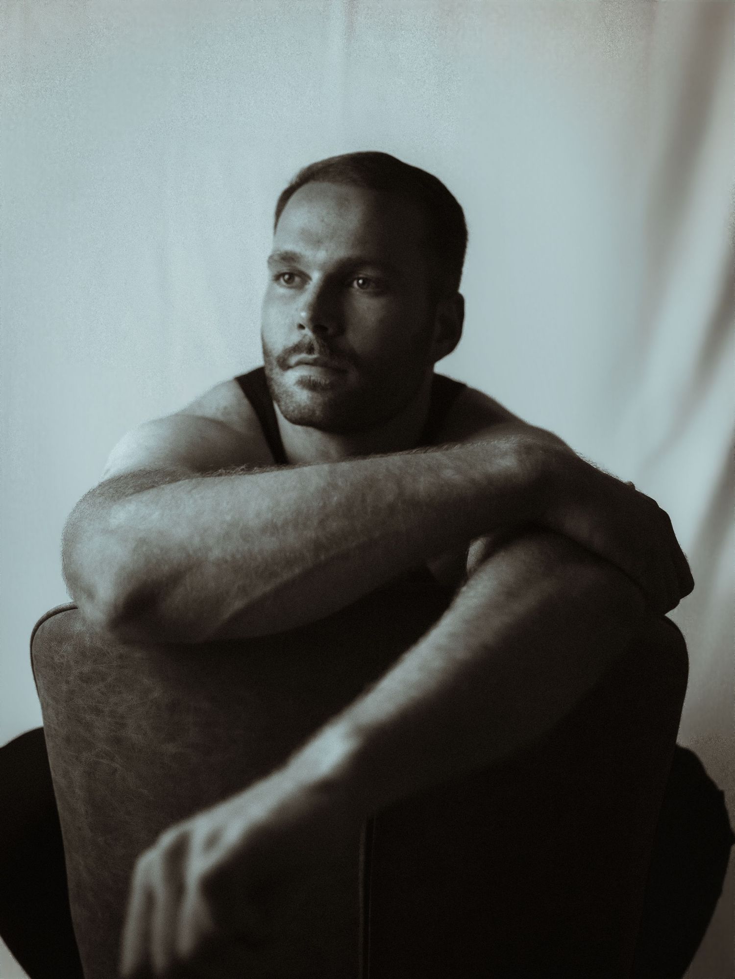 Moody black and white portrait of a bearded man sitting pensively with arms crossed over knees.
