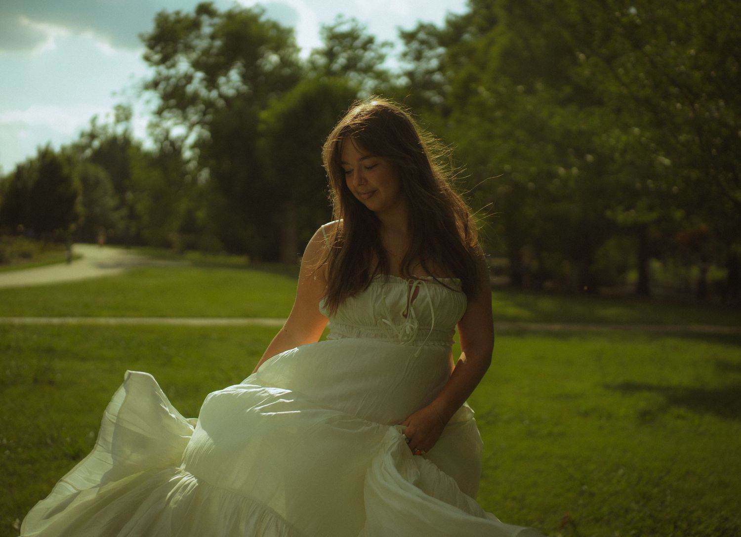 Young woman in flowing white wedding dress standing gracefully in a lush green park on a sunny day.