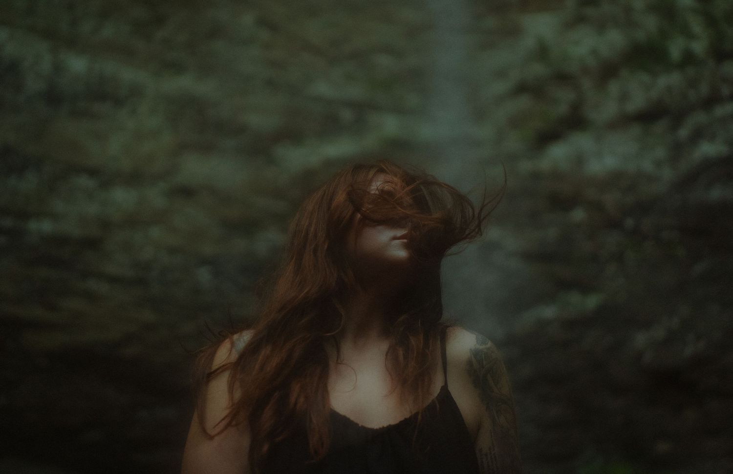 Woman with long red hair blowing across her face standing in a dark moody forest setting.