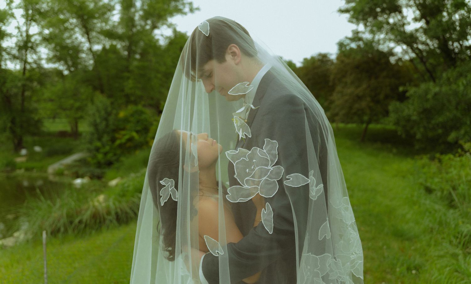 Bride and groom sharing an intimate moment under a floral embroidered wedding veil in a lush green outdoor setting.