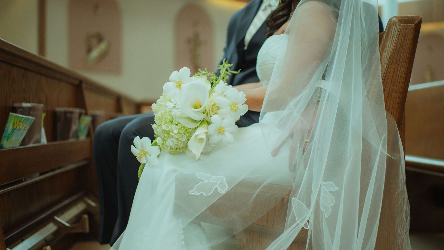 Bride in white wedding dress holding bouquet of white flowers while sitting in church pew beside groom.