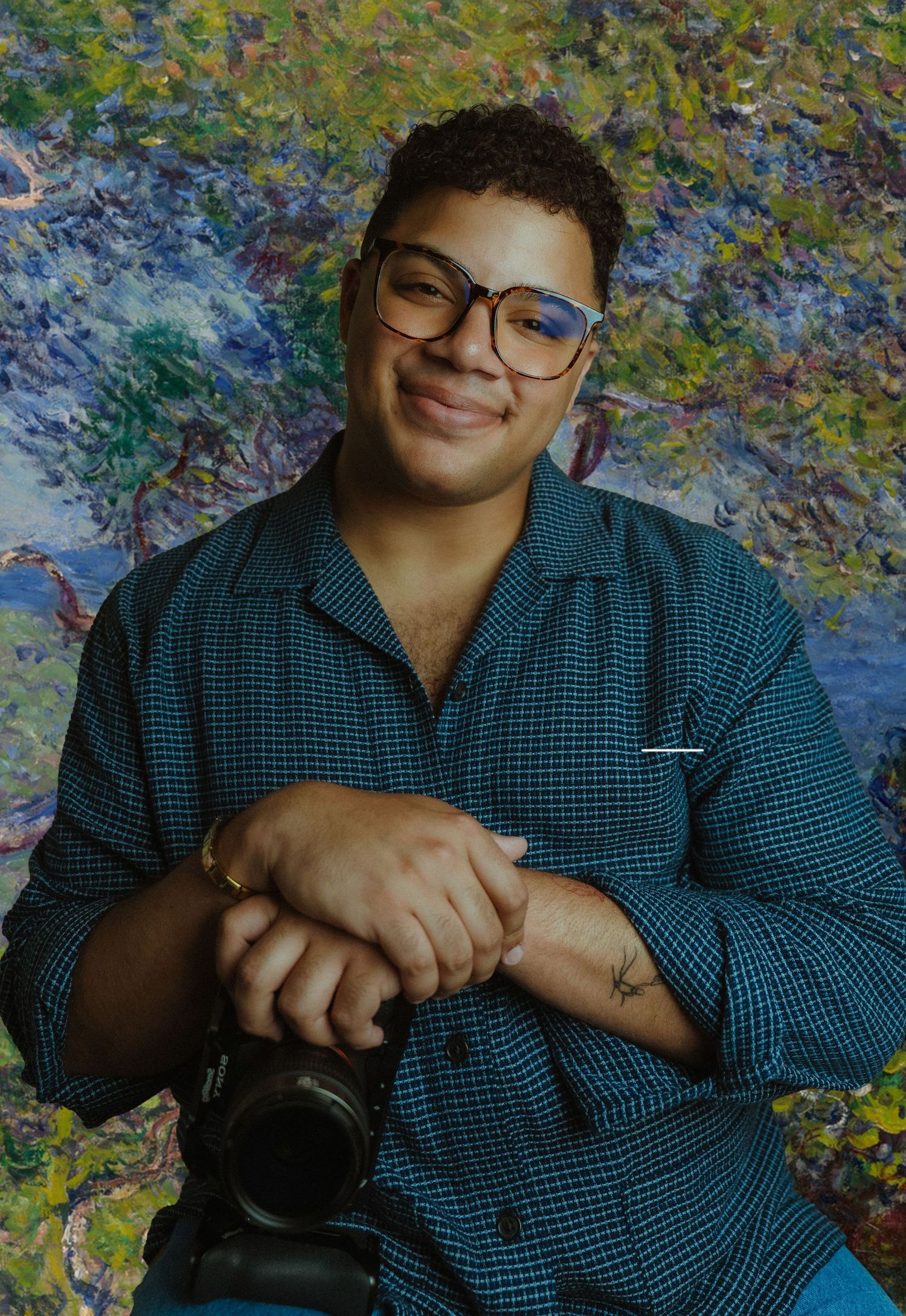 Person with glasses holding a camera, smiling in front of a colorful impressionist painting backdrop.