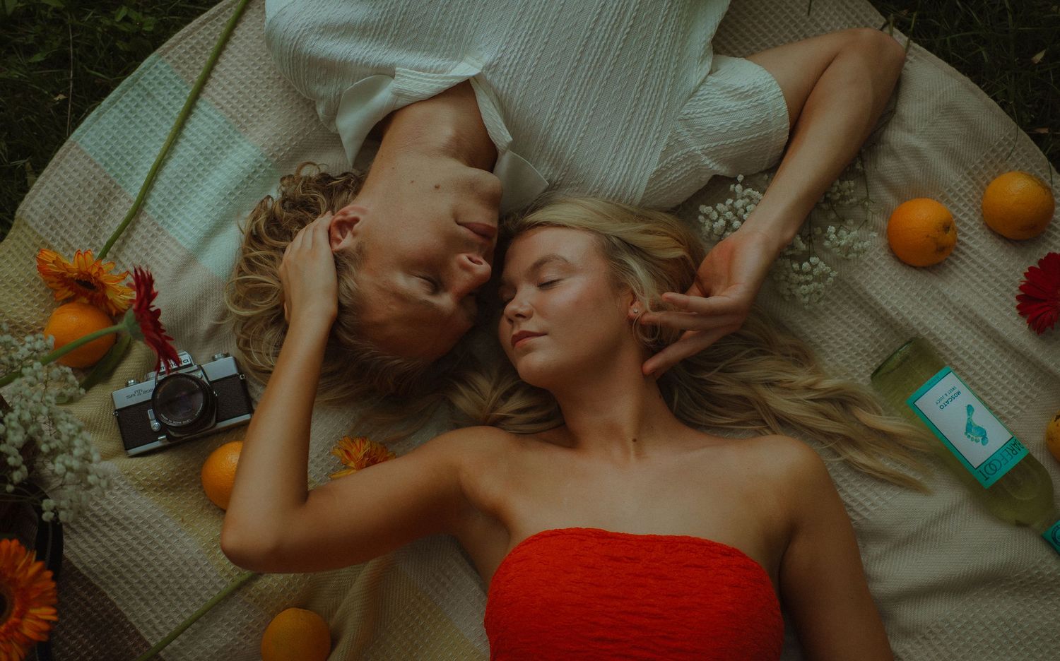 Two women lying on a blanket surrounded by oranges and flowers, sharing a peaceful moment outdoors.
