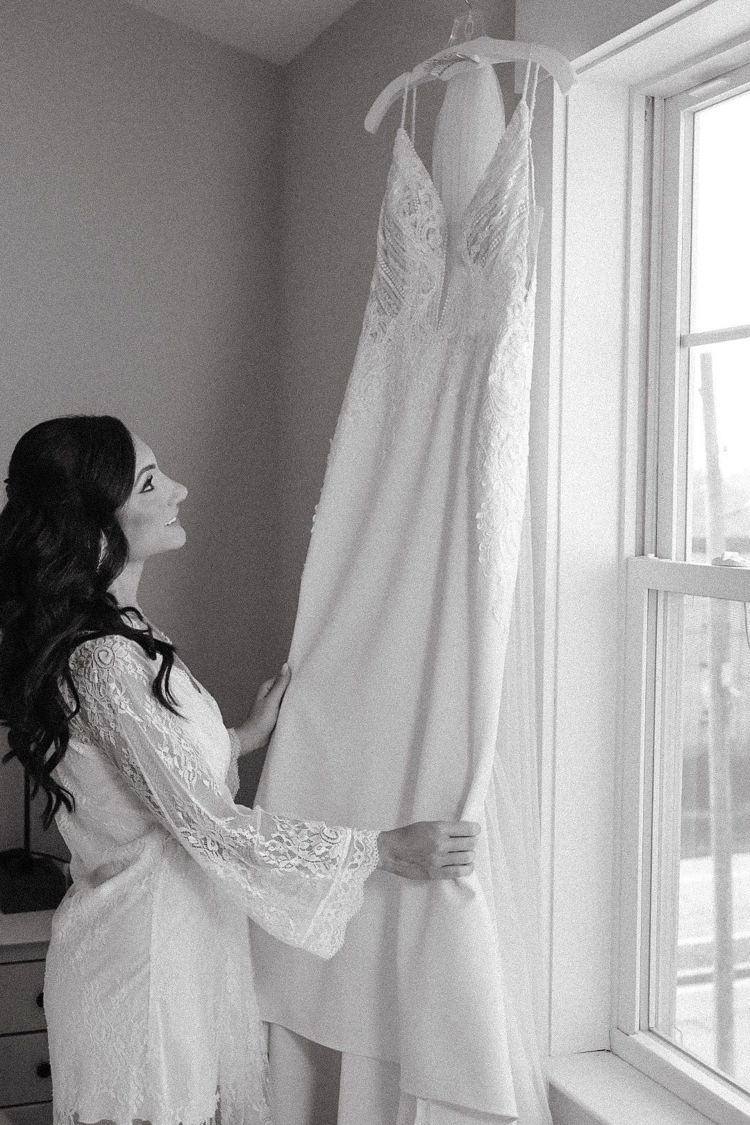 A soft, intimate portrait of a bride in a lace robe gazing at her wedding gown hanging by a window, reflecting quiet anticipation.