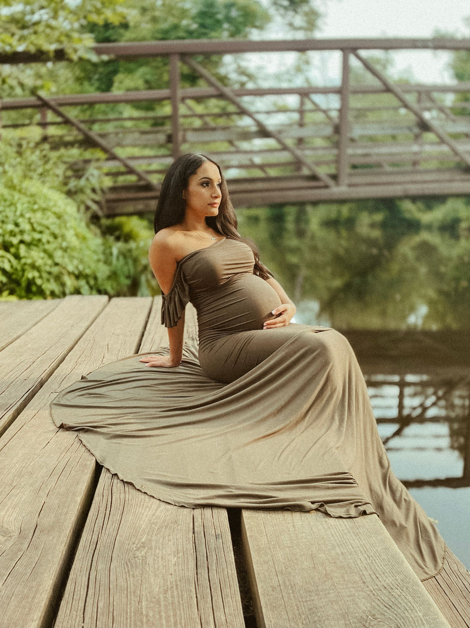 Intimate profile portrait of a pregnant woman in an earth-toned dress, captured against a lush greenery and water backdrop.