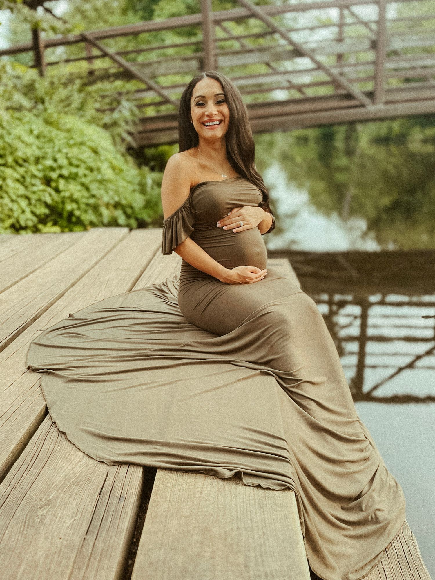Expectant mother in a flowing olive-toned maternity gown sitting gracefully on a rustic wooden bridge during a maternity session.