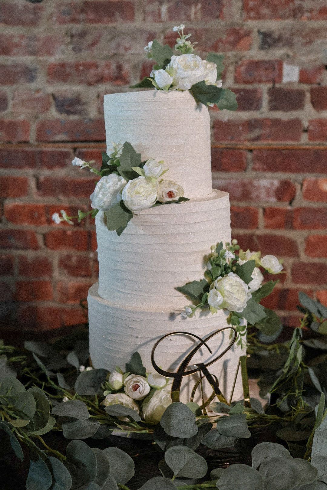 A classic three-tier white wedding cake adorned with fresh white roses and greenery, displayed against a rustic red brick wall.