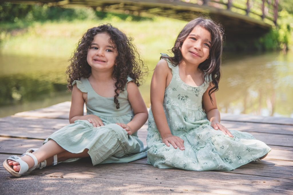 Two young girls in matching floral dresses sitting by a peaceful pond, reflecting a timeless and artful childhood portrait.