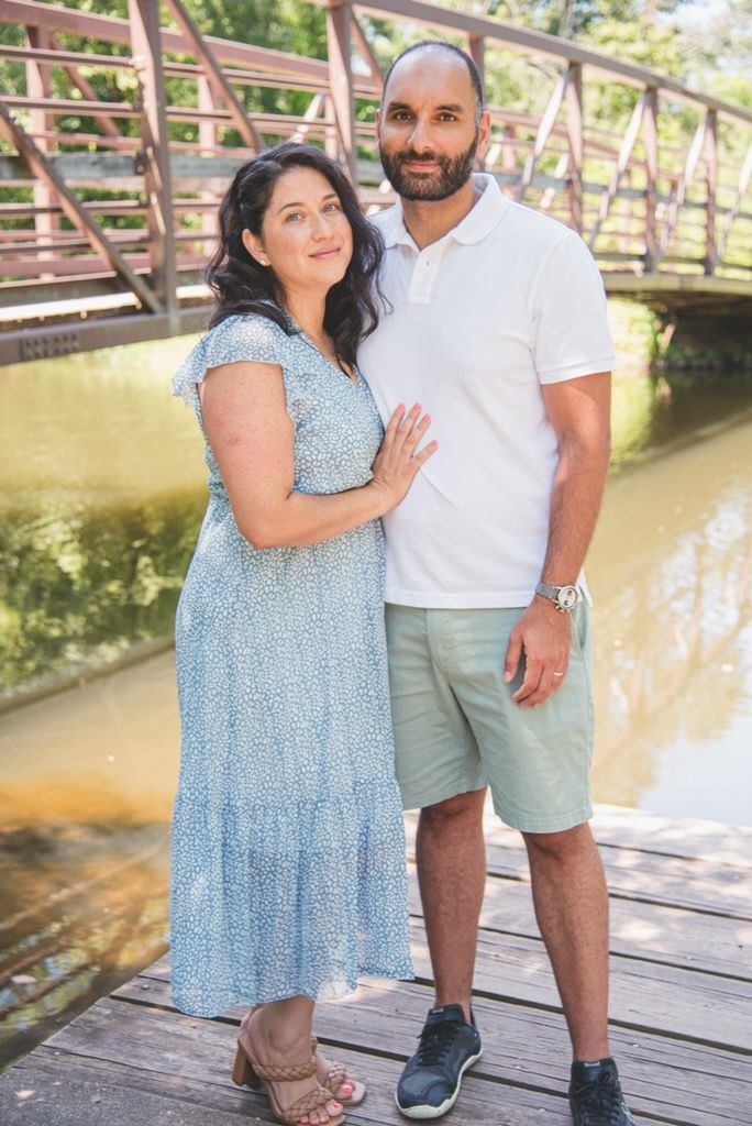 Full-length portrait of a couple standing on a wooden bridge over a peaceful pond, reflecting a timeless and elegant aesthetic.