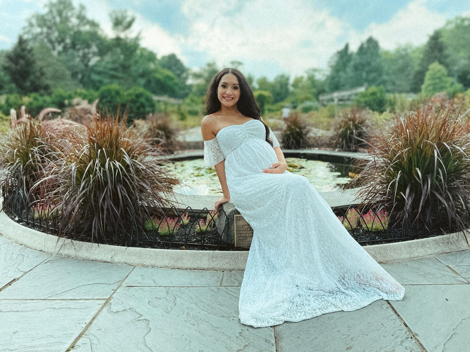 Expectant mother in an elegant off-the-shoulder white lace gown posing gracefully by a stone garden pond.