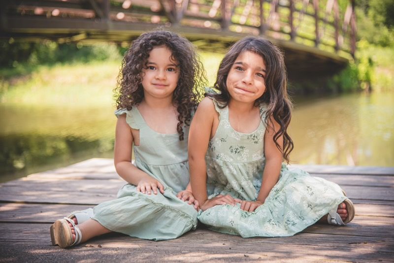 Two young sisters in matching sage green dresses sitting together on a wooden bridge, showcasing timeless and soulful childhood photography.