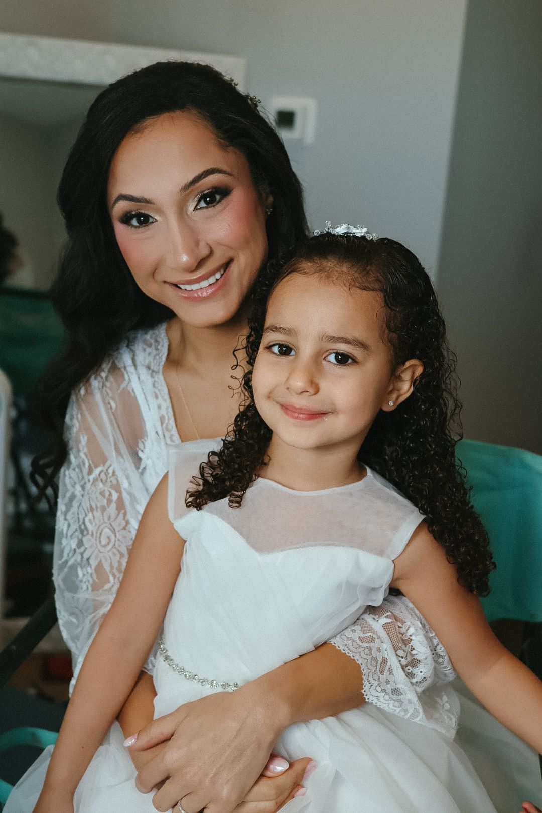 A smiling bride sitting with a young flower girl, showcasing the sweet and joyful connection shared during the wedding preparations.