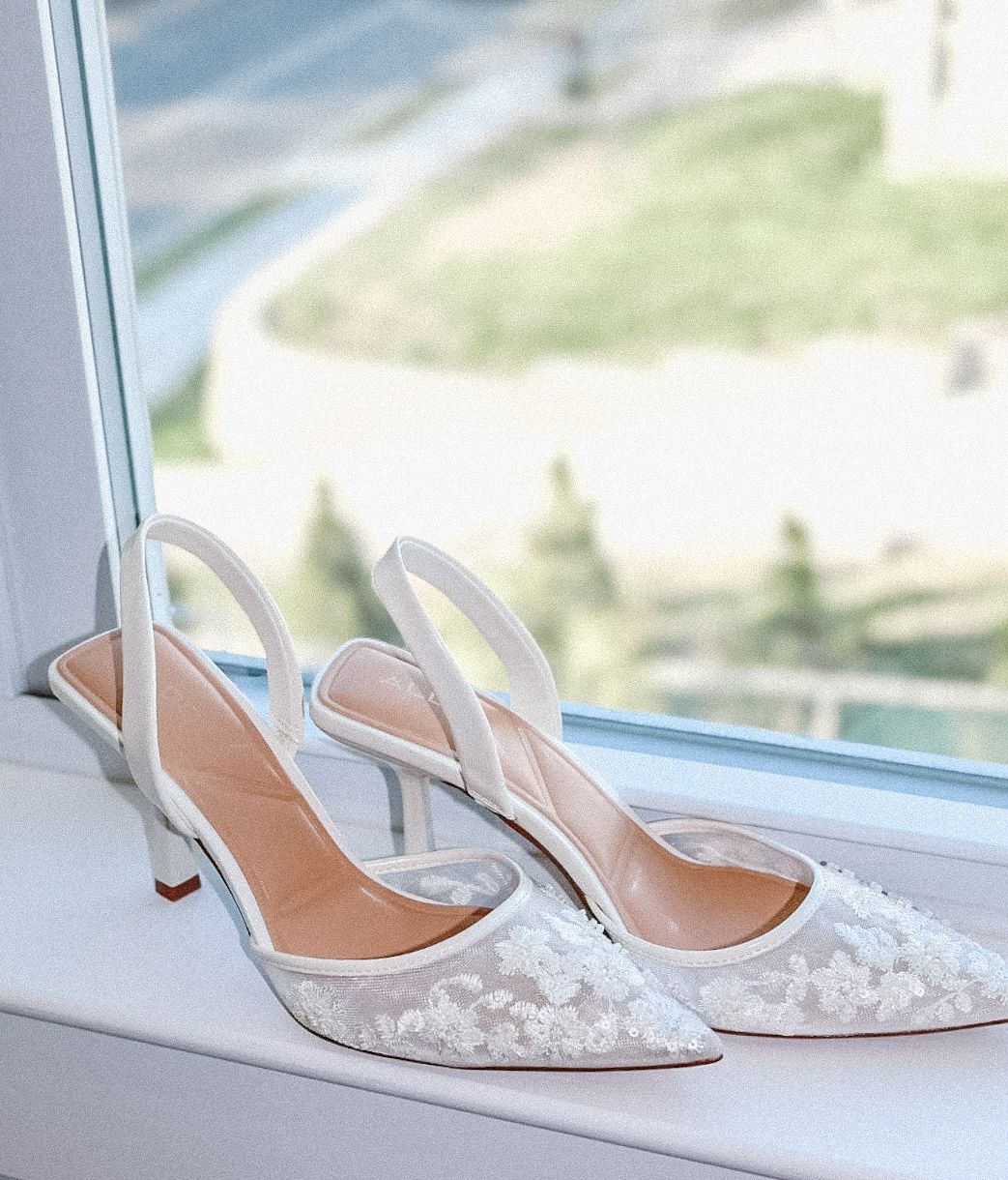 Elegant white lace bridal heels placed on a window sill in soft, natural light, reflecting the beautiful details of the wedding morning prep