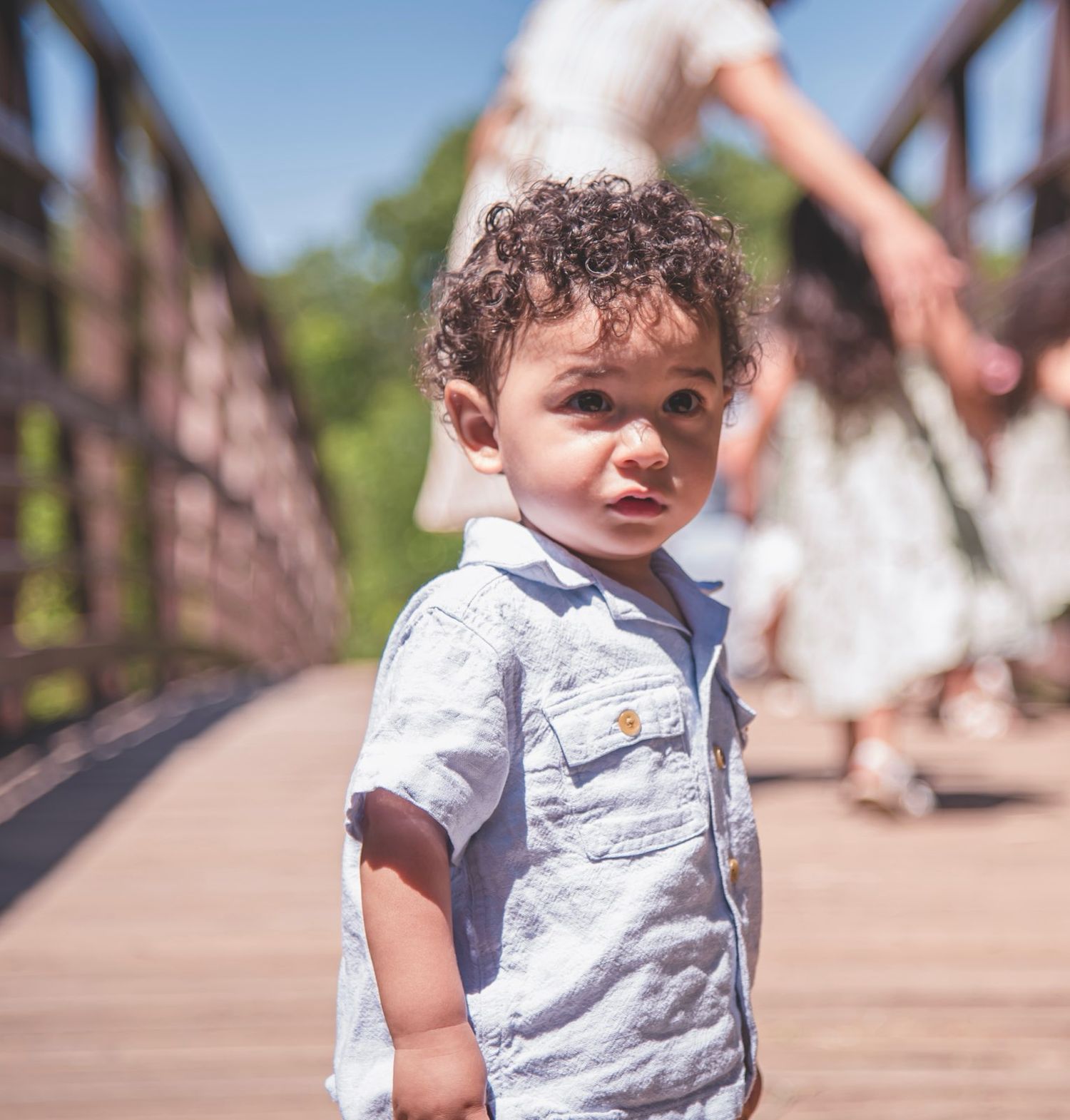 A candid close-up of a toddler boy in a blue button-down shirt walking across a rustic bridge during a family portrait session.