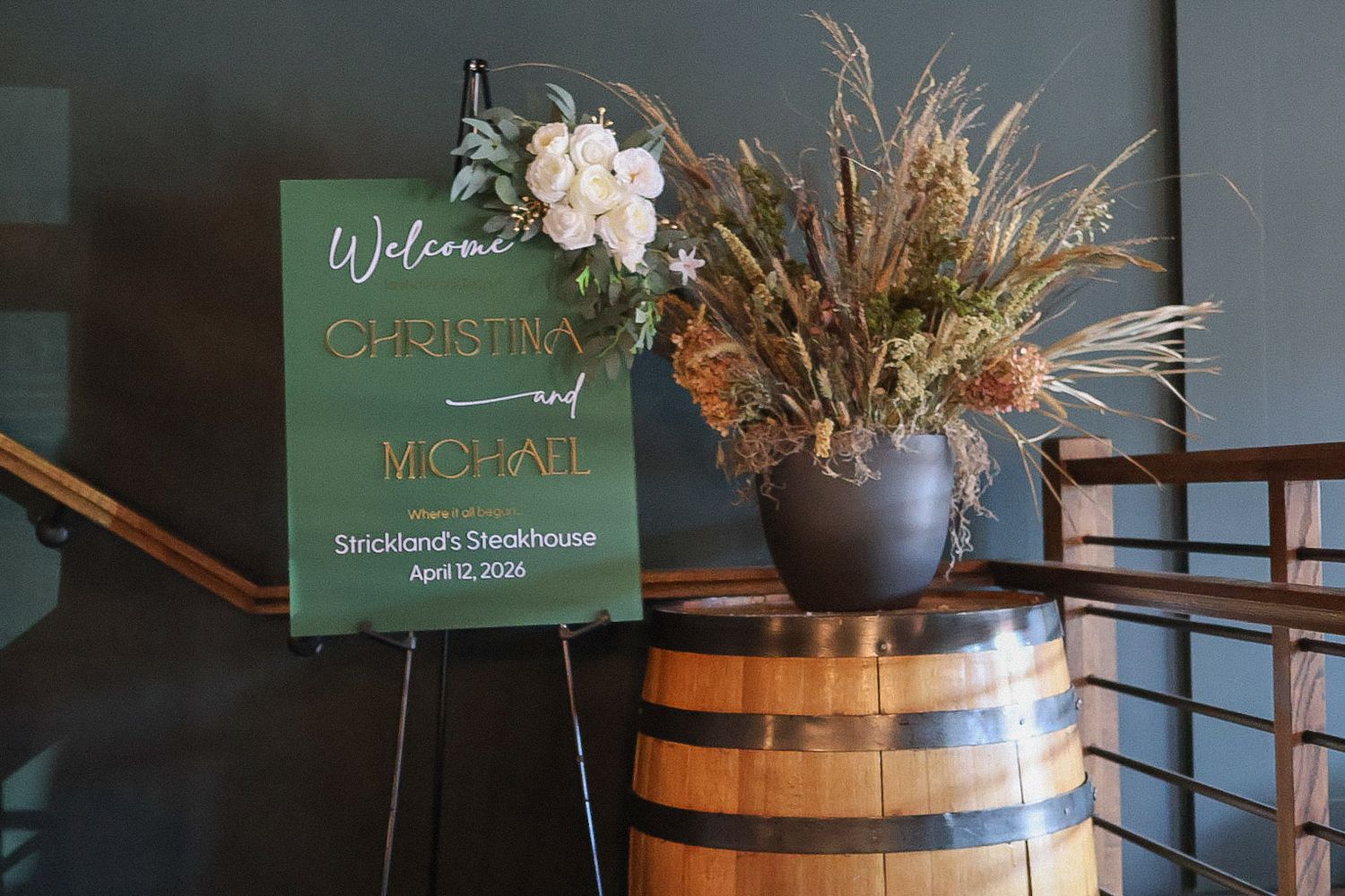 A wooden wedding welcome sign decorated with white flowers and dried grass, set against a rustic barrel at a reception entrance.