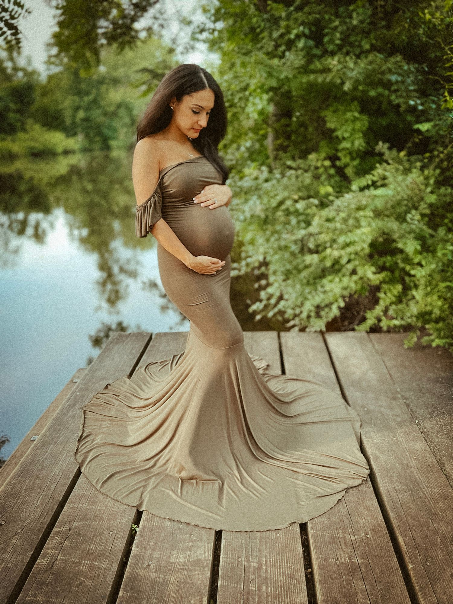 Serene maternity session featuring a woman in a sage green dress standing by a pond, reflecting a sense of peace and natural beauty.