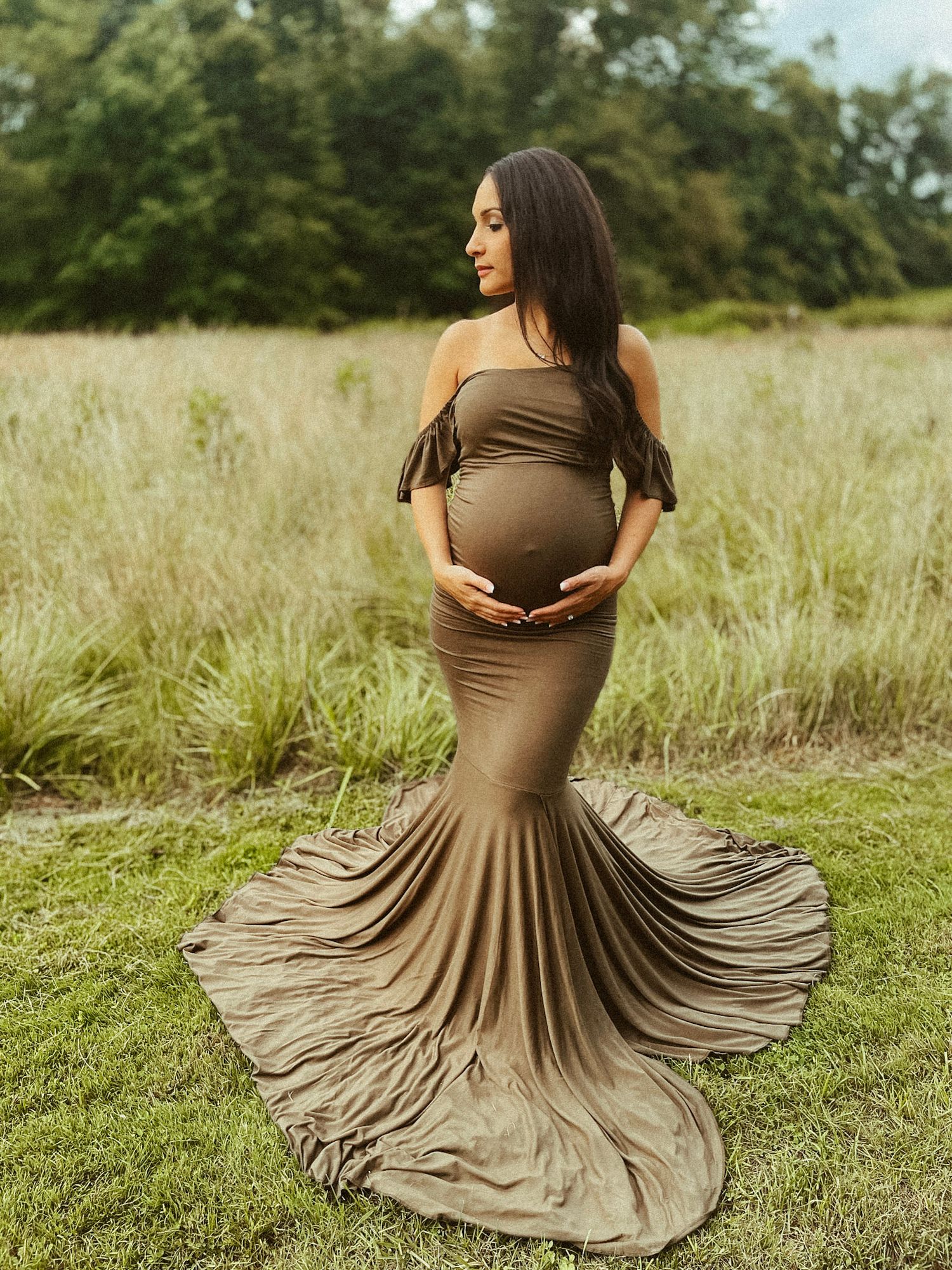Beautiful maternity portrait of a woman in a flowing earth-toned dress standing in a serene, sun-drenched field.