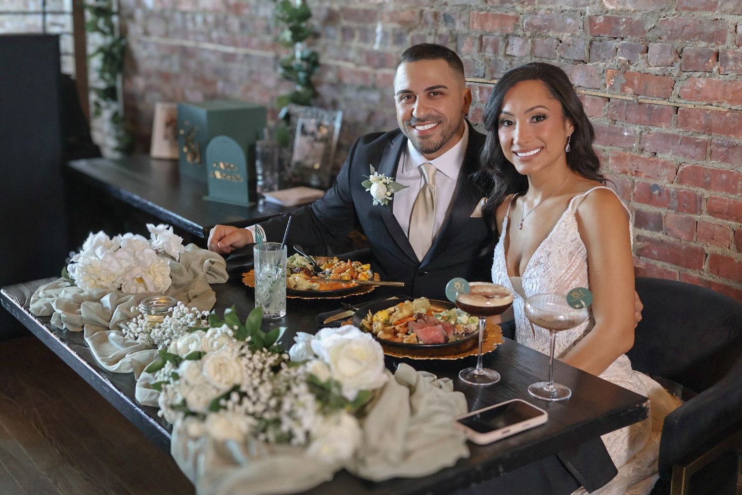 A joyful bride and groom sitting at their decorated reception table, sharing a meal and a smile in front of a rustic brick wall.