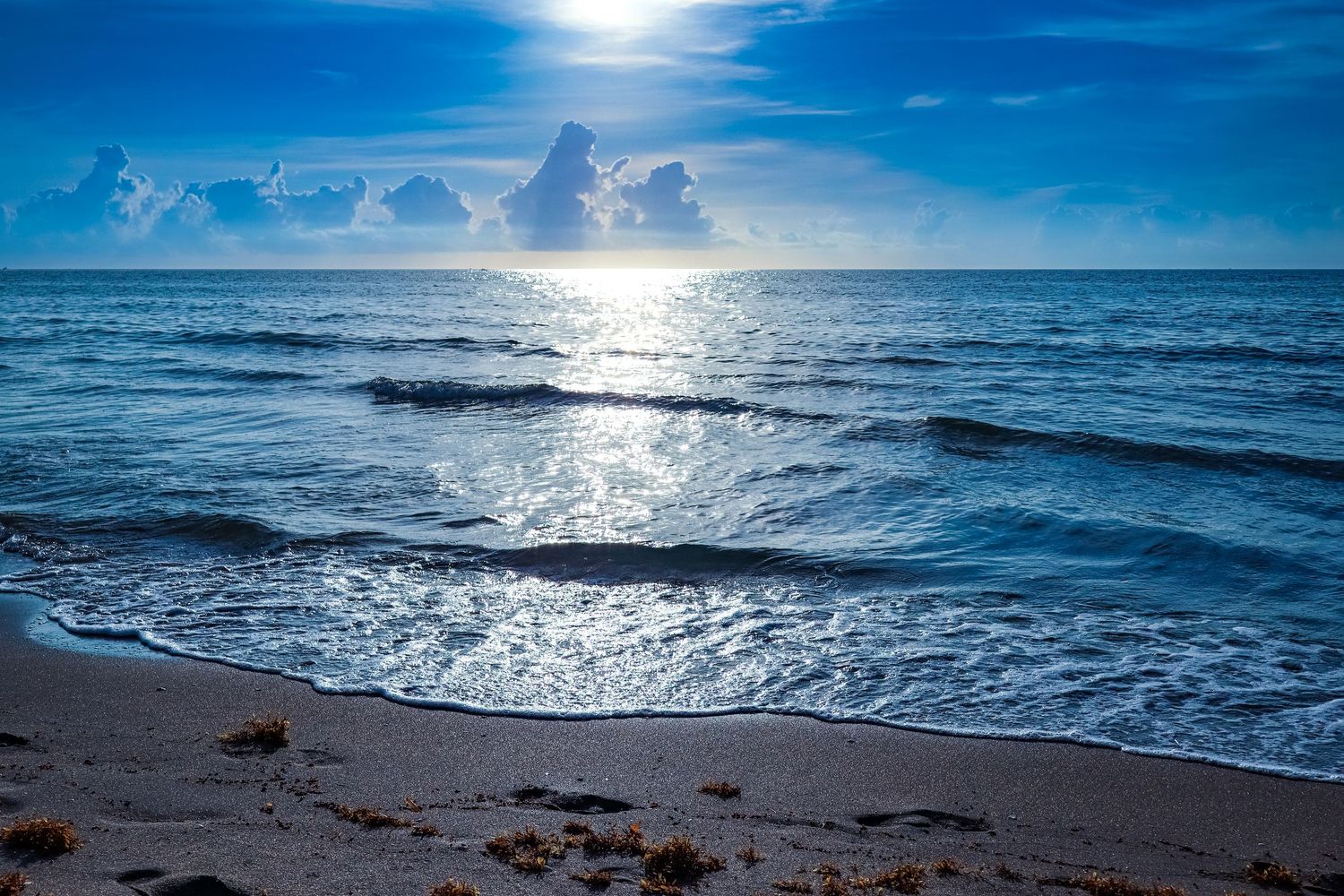 Serene ocean beach scene with gentle waves, white sand, and dramatic blue sky with clouds at sunset.