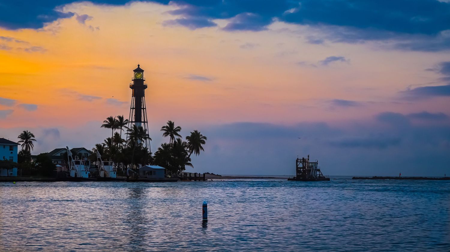 Hillsboro Inlet Lighthouse silhouetted against a vibrant orange and purple sunset sky with palm trees in Florida.