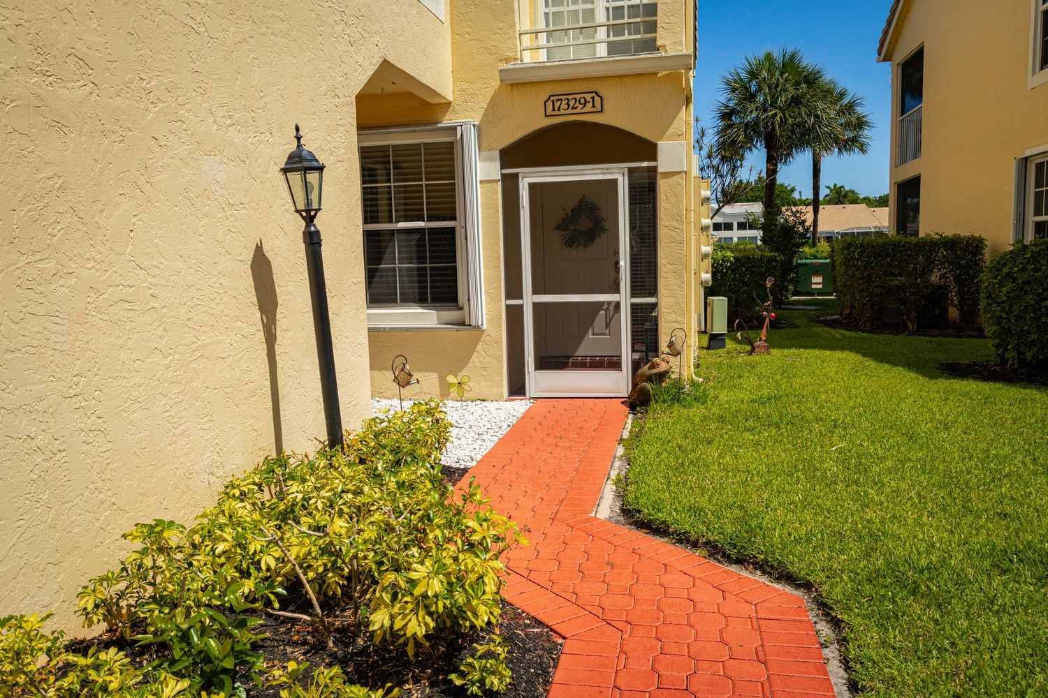 Florida home entrance with red brick pathway, manicured shrubs, screen door, and lush green lawn.