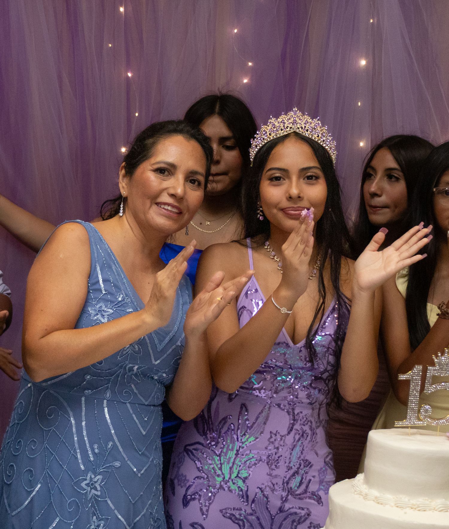 Young woman wearing tiara and purple gown celebrates quinceañera with family beside white cake.
