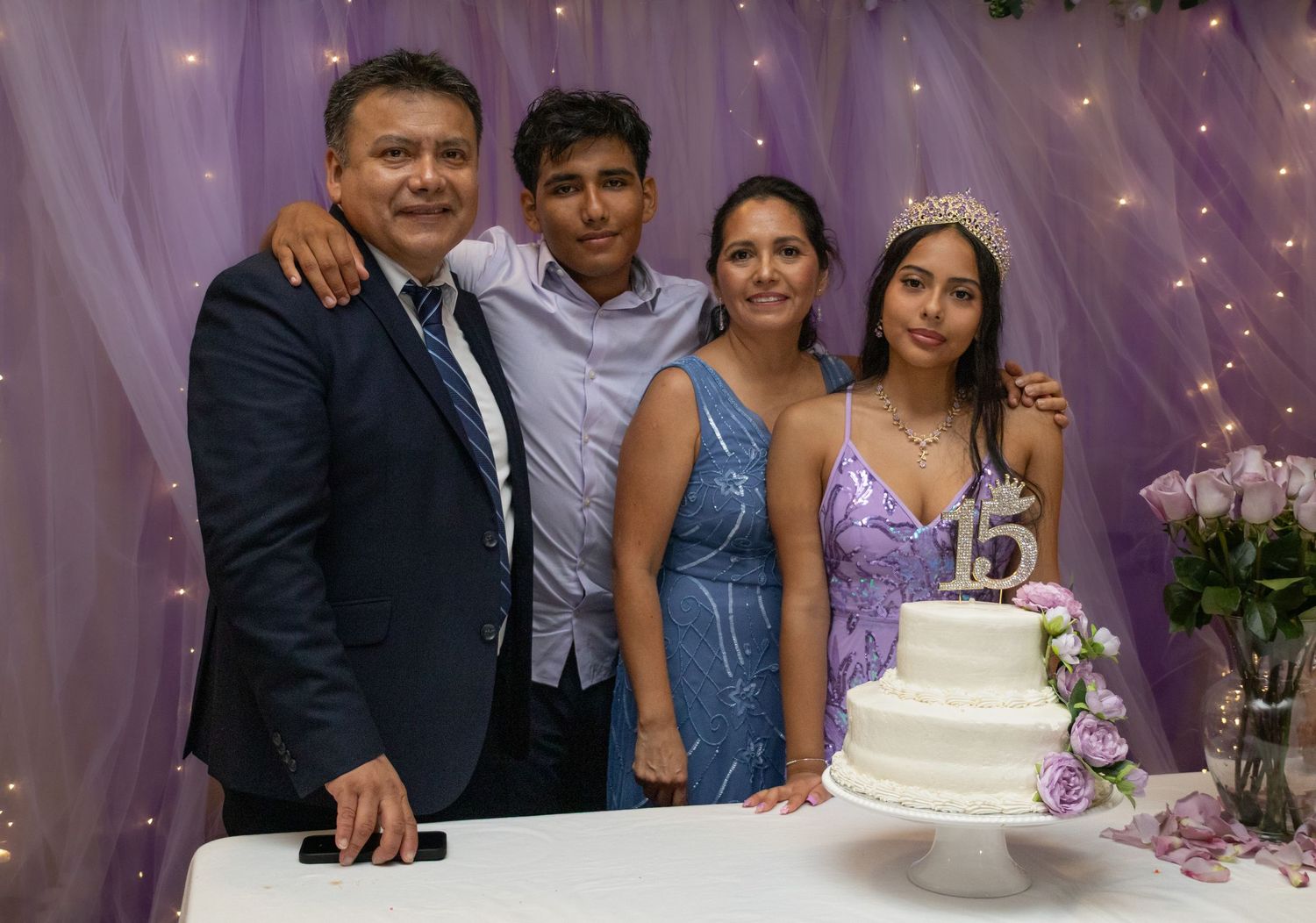 Family of four posing together at a quinceañera celebration with a white cake displaying the number 15.