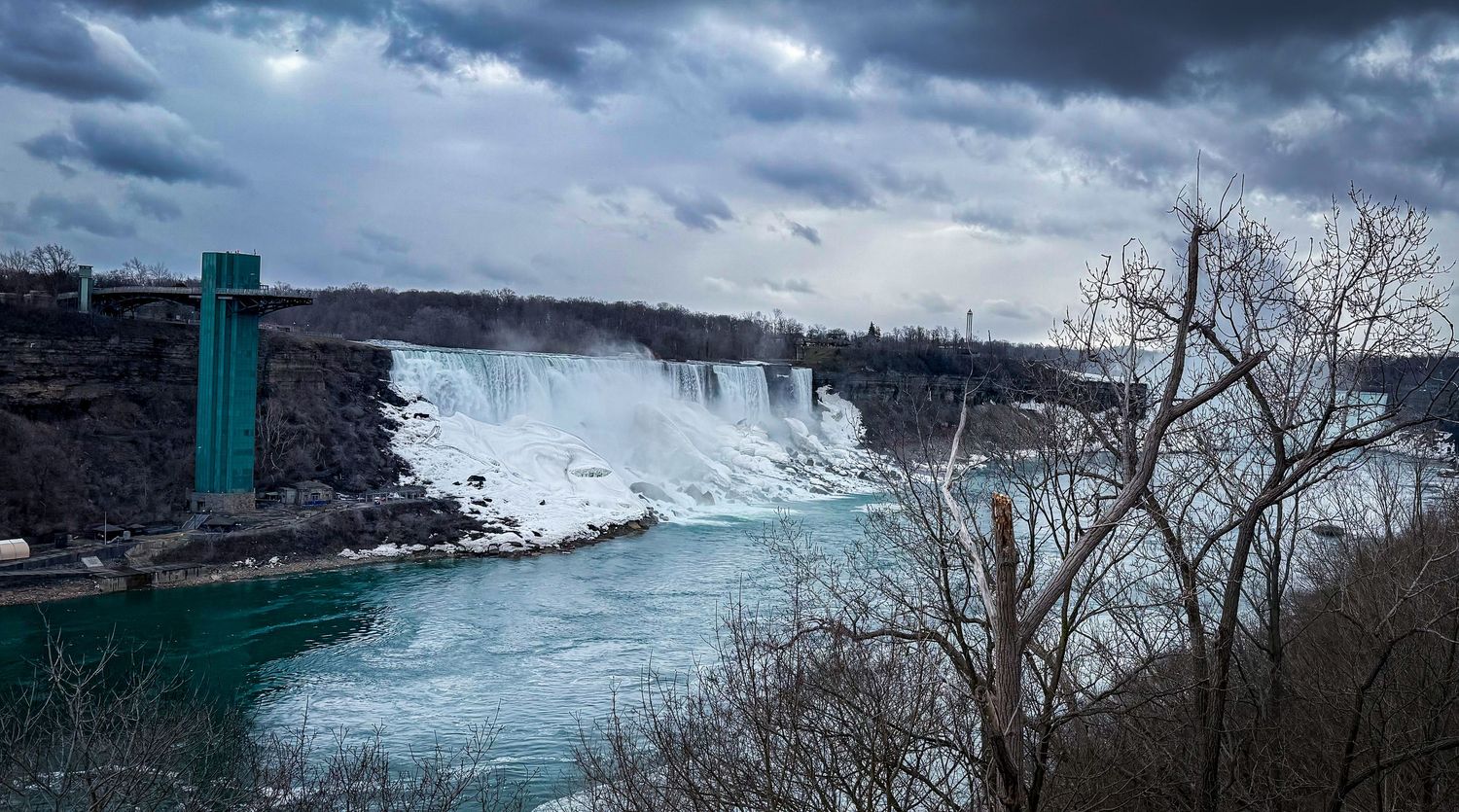 Niagara Falls panoramic winter view with icy blue waters and snow-covered rocks under dramatic cloudy sky.