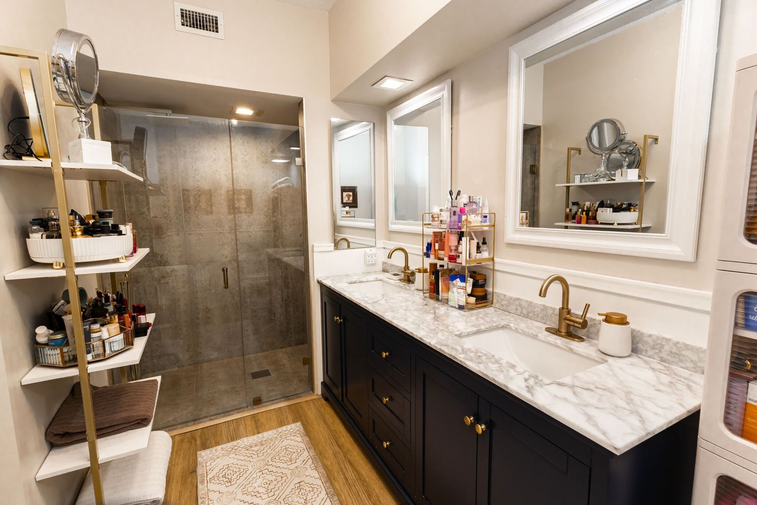 Elegant bathroom with dark navy double vanity, marble countertop, gold fixtures, glass shower, and open shelving.