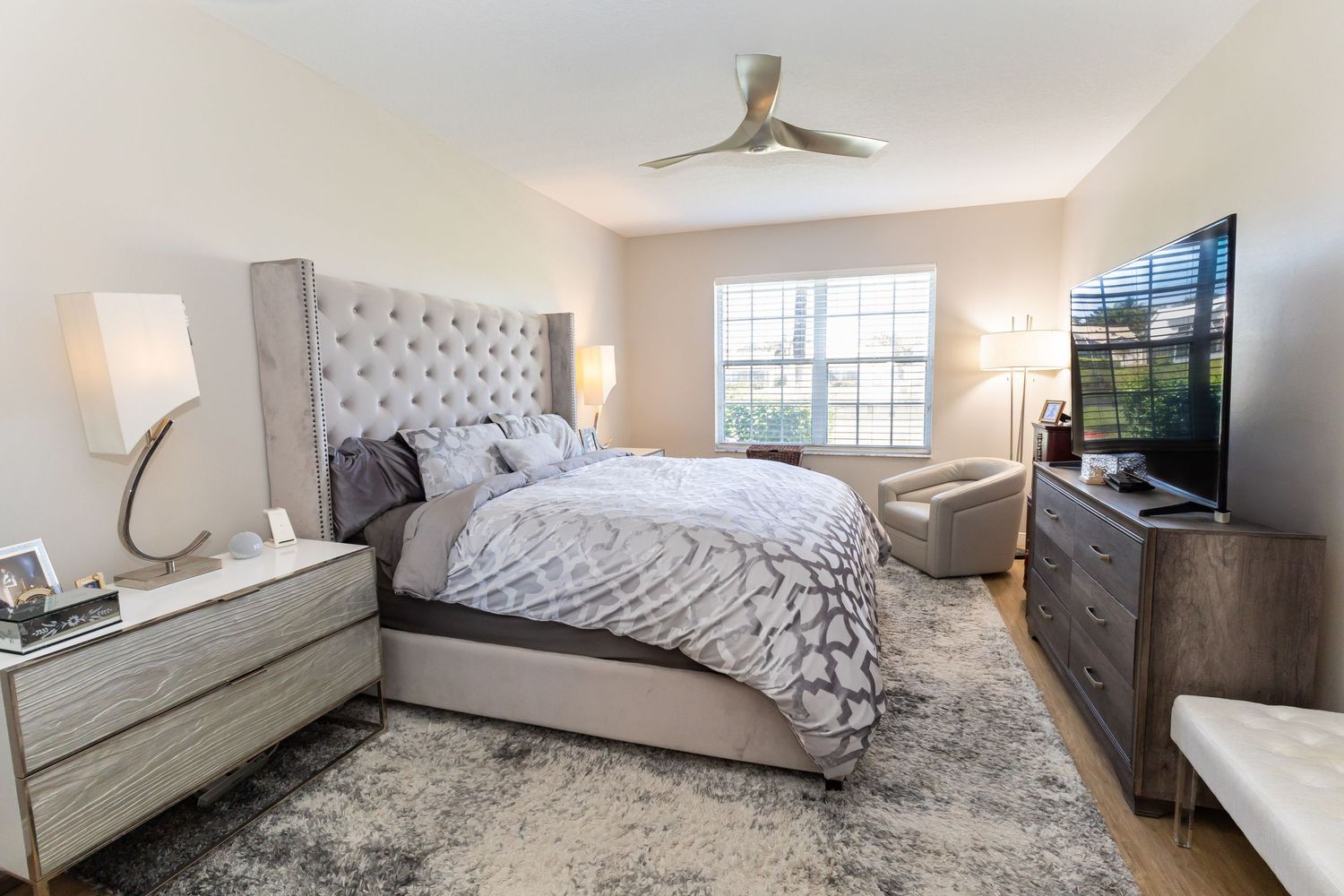 Spacious master bedroom with tufted gray headboard, ceiling fan, large TV, and neutral modern decor.